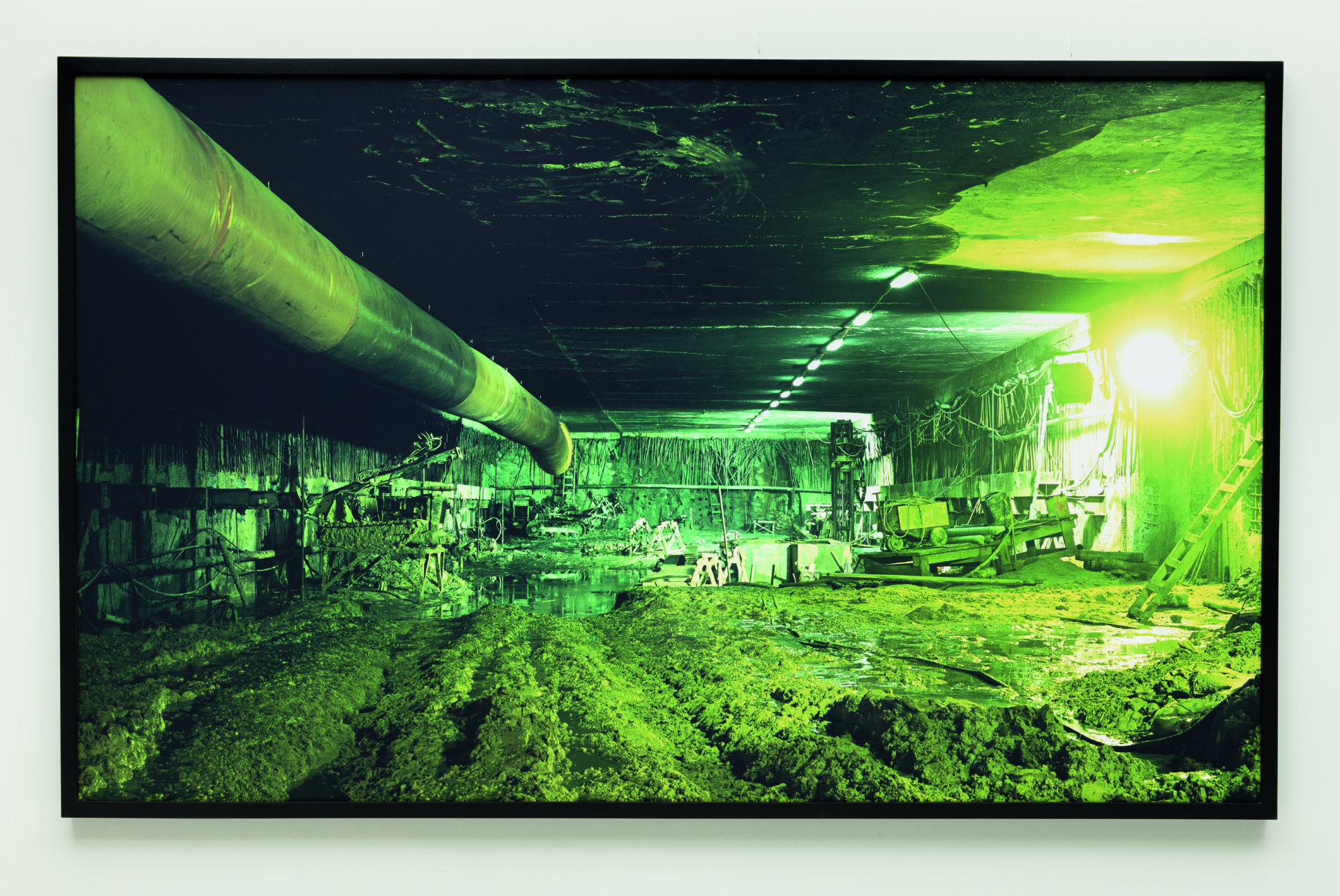 Descrição da imagem: Fotografia de um ambiente subterrâneo iluminado por luzes artificiais com reflexo esverdeado. À esquerda, um grande tubo percorre o teto. No chão, detritos estão misturados com água. À direita, há uma luz muito forte e uma escada encostada na parede. Ao fundo, há máquinas, equipamentos industriais e estruturas metálicas. A moldura é fina e preta. Fim da descrição. 

