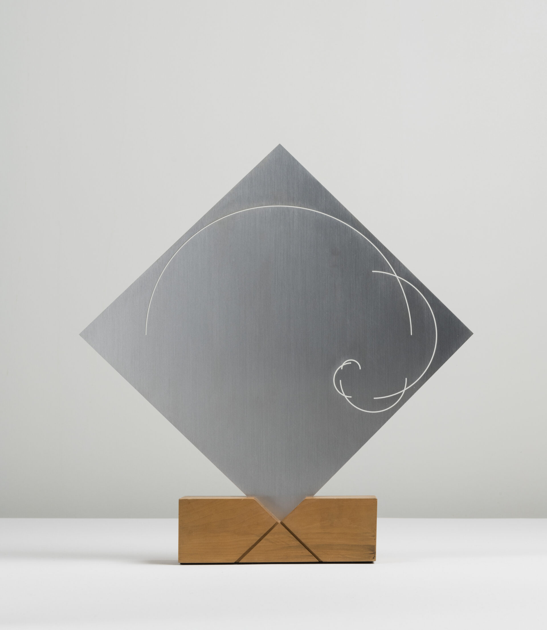 Descrição da imagem: A escultura é composta por uma placa quadrada de alumínio anodizado — um tratamento que forma uma camada protetora na superfície do metal — posicionada em diagonal, com um dos vértices voltado para cima. 
Na superfície do metal, traços curvos gravados se conectam entre si. A peça metálica está inserida no encaixe em “V” de um bloco retangular de madeira clara. Fim da descrição.
