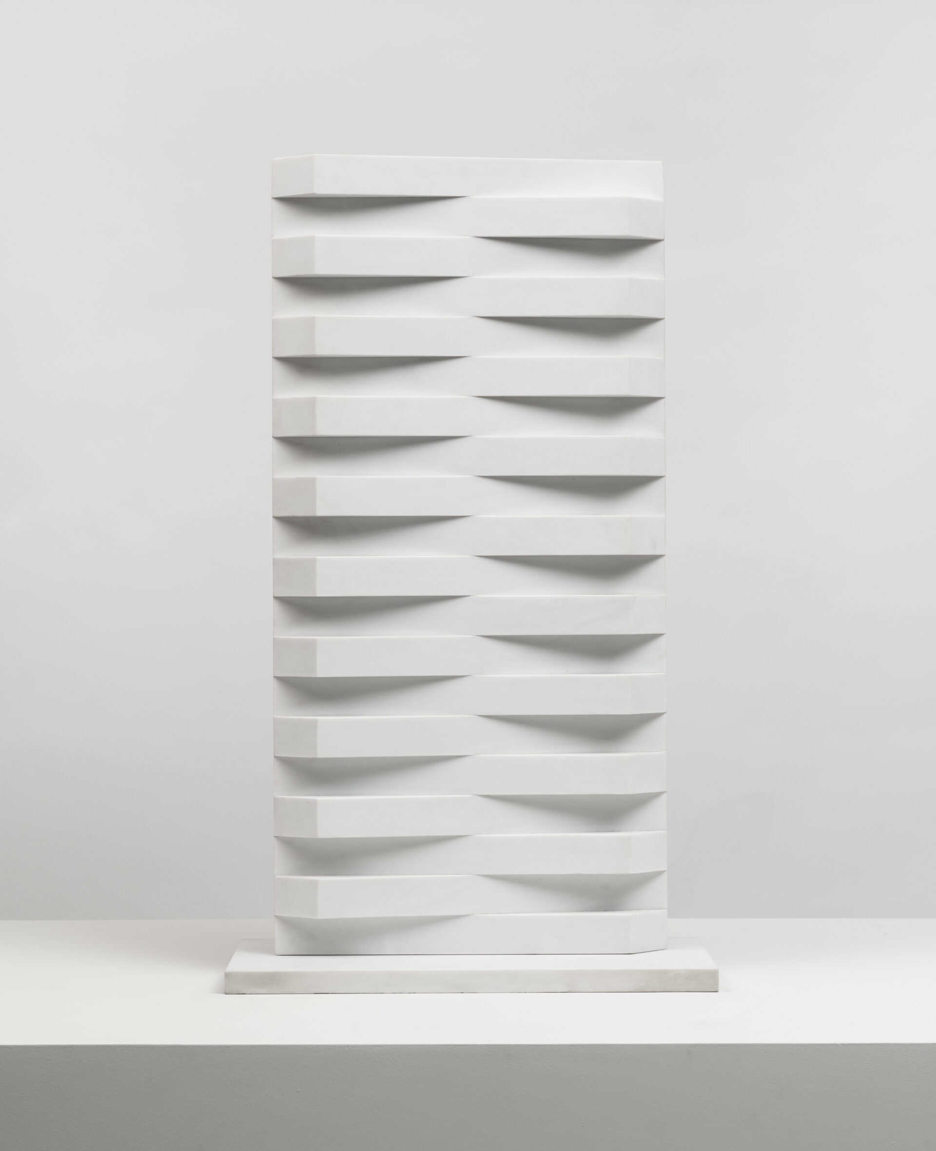 Descrição da imagem: Escultura de mármore de Carrara, branca e em formato retangular alongado, apoiada sobre uma base plana e retangular, ligeiramente mais larga. Ao longo da escultura, repete-se um padrão intercalado de trapézios em alto e baixo relevo: de um lado, os trapézios se projetam para fora, e, na mesma linha, o lado oposto está recuado. Fim da descrição. 
