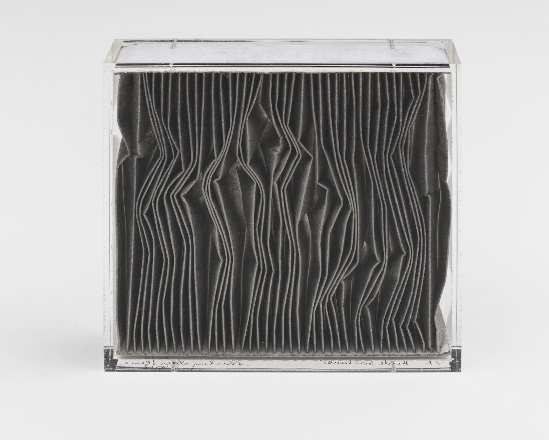 Descrição da imagem: Escultura de uma caixa acrílica retangular transparente. Dentro da caixa, há um filtro de ar automotivo cinza-escuro. Ele é formado por pregas verticais, com ondulações irregulares centralizadas. Fim da descrição. 
