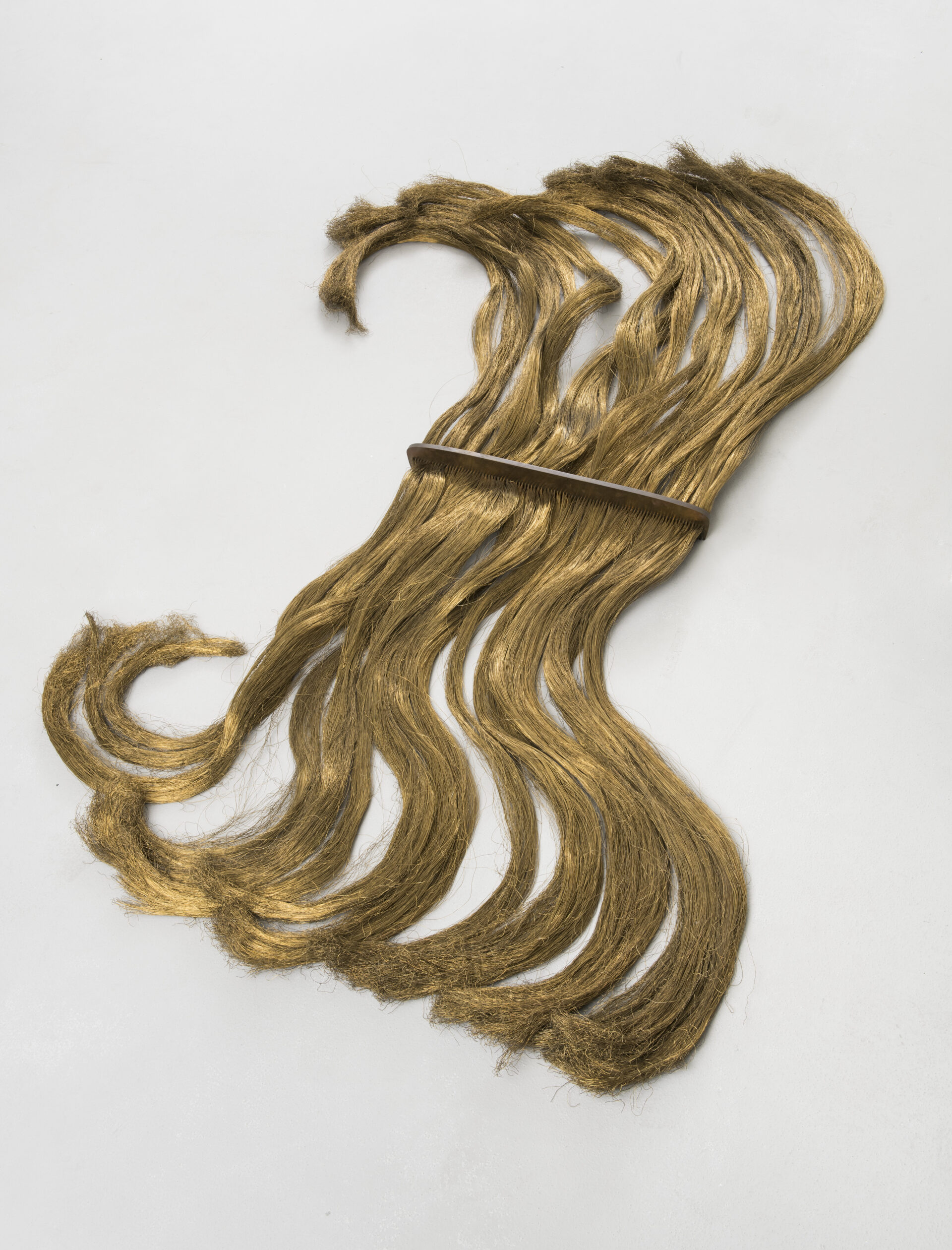 Descrição da imagem: Obra composta por grandes mechas de cabelo. As mechas são loiras, longas e lisas e estão presas no meio por um pente de cobre. Fim da descrição. 
