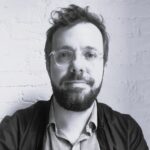 Imagem. Início da descrição de imagem: Retrato frontal em preto e branco de um homem de pele clara, com cabelos curtos e desalinhados em um topete no topo da cabeça. Ele tem uma barba cheia e bem aparada. Ele usa óculos de grau com armação transparente e circular, e veste uma camisa de tom médio sob uma jaqueta escura aberta. O homem olha diretamente para a câmera com uma expressão neutra e serena, posicionado à frente de uma parede de tijolos pintada de branco. Fim da descrição de imagem.