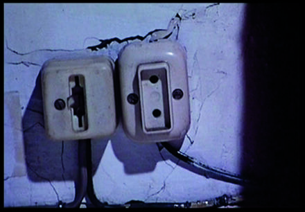 Descrição da imagem: duas tomadas estão fixadas lado a lado em uma parede branca com rachaduras. Fios saem da parte inferior das tomadas. Fim da descrição. 
