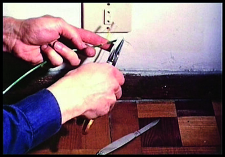 Descrição da imagem: mãos seguram um alicate e um fio, perto de uma tomada no rodapé de uma parede branca. No chão de pequenos azulejos marrons, há uma pequena faca. Fim da descrição. 

