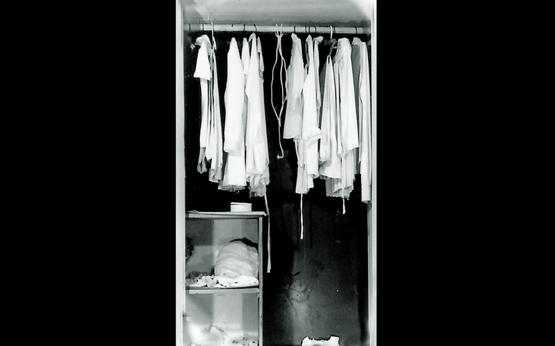Descrição da imagem: roupas claras penduradas em cabides dentro de um guarda-roupa. Dentro dele, no canto inferior esquerdo, há duas caixas, uma em cima da outra, com objetos dentro. Fim da descrição. 