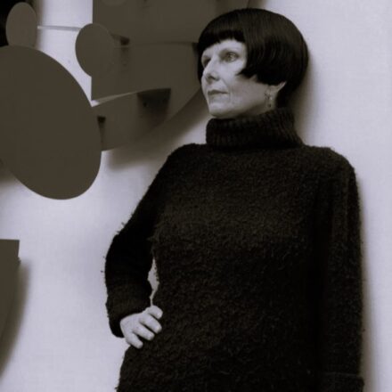 A imagem é um retrato em preto e branco de uma mulher de meia-idade ou idosa, de pele clara, posicionada de frente para a câmera, mas olhando para sua esquerda. Ela tem cabelo preto liso, cortado em estilo "chanel" ou tigela, com franja reta. Sua pele é pálida e seus olhos são claros. A mulher veste um suéter de gola rolê, de tricô grosso e escuro. Sua mão direita está apoiada na cintura, e seu braço esquerdo está estendido para baixo. O fundo é uma parede clara e lisa. Atrás e à esquerda da mulher, há uma obra de arte abstrata composta por formas geométricas circulares e angulares em tons escuros, que parecem estar fixadas na parede. A iluminação é suave, com a luz vindo da esquerda, criando um leve contraste de luz e sombra no rosto e na obra.