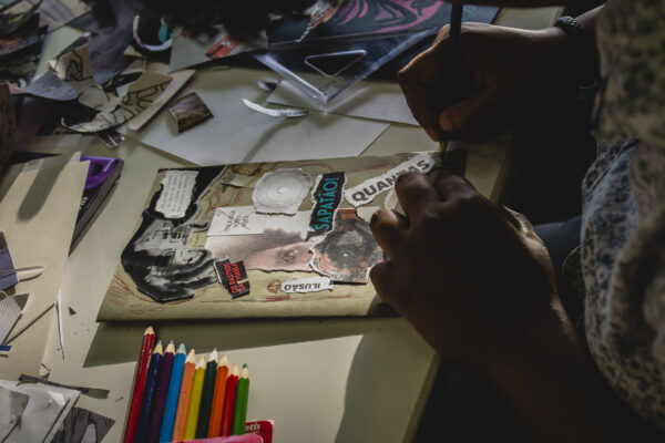 Descrição da imagem: uma foto com iluminação baixa e foco na atividade de colagem e criação de zine. As mãos escuras de uma pessoa estão usando um instrumento pontiagudo (lápis ou estilete) para trabalhar em uma colagem que está sendo montada sobre um papel em tom kraft ou bege. A colagem é composta por recortes de revistas, palavras e imagens em preto e branco com temática urbana ou subversiva (palavras visíveis incluem 