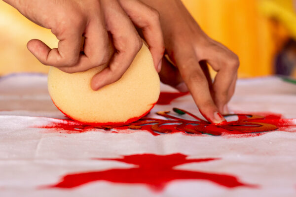 Descrição da imagem: um close-up mostrando as mãos de uma pessoa realizando uma técnica de estamparia ou stencil. As mãos estão posicionadas sobre um tecido branco que já possui manchas de tinta vermelha. A mão direita segura uma esponja redonda e bege (laranja-claro) cuja parte inferior está saturada de tinta vermelha. A esponja está sendo pressionada sobre um objeto de contorno em formato de folha ou flor. A mão esquerda apoia ou guia a estamparia. O fundo está fora de foco, em tons de amarelo e laranja brilhantes. Fim da descrição.