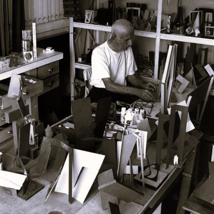 A imagem é uma fotografia em preto e branco que retrata um homem idoso em seu ateliê, trabalhando com diversas peças de arte abstrata. O homem, que é calvo, veste uma camiseta branca simples de manga curta e calças escuras. Ele está sentado em uma cadeira, curvado sobre uma mesa de trabalho no centro da imagem. Suas mãos estão ocupadas manuseando pequenos objetos ou materiais na superfície da mesa. Ele está olhando para baixo, concentrado na sua tarefa. A mesa de trabalho está repleta de ferramentas e materiais, incluindo tubos e frascos de cola ou tinta, e várias pequenas esculturas ou modelos abstratos. Ao redor do homem e da mesa, o ateliê está completamente preenchido com inúmeras obras de arte e materiais. Há estantes ao fundo e à direita, cheias de objetos geométricos, formas recortadas e pequenas esculturas em diferentes tons e materiais. Muitos desses objetos são abstratos, com ângulos retos e planos. No chão, ao redor da mesa, mais peças de arte e recortes estão espalhados, criando uma sensação de um espaço criativo e produtivo. A iluminação é difusa, mas as sombras acentuam a tridimensionalidade dos objetos e a profundidade do ateliê.