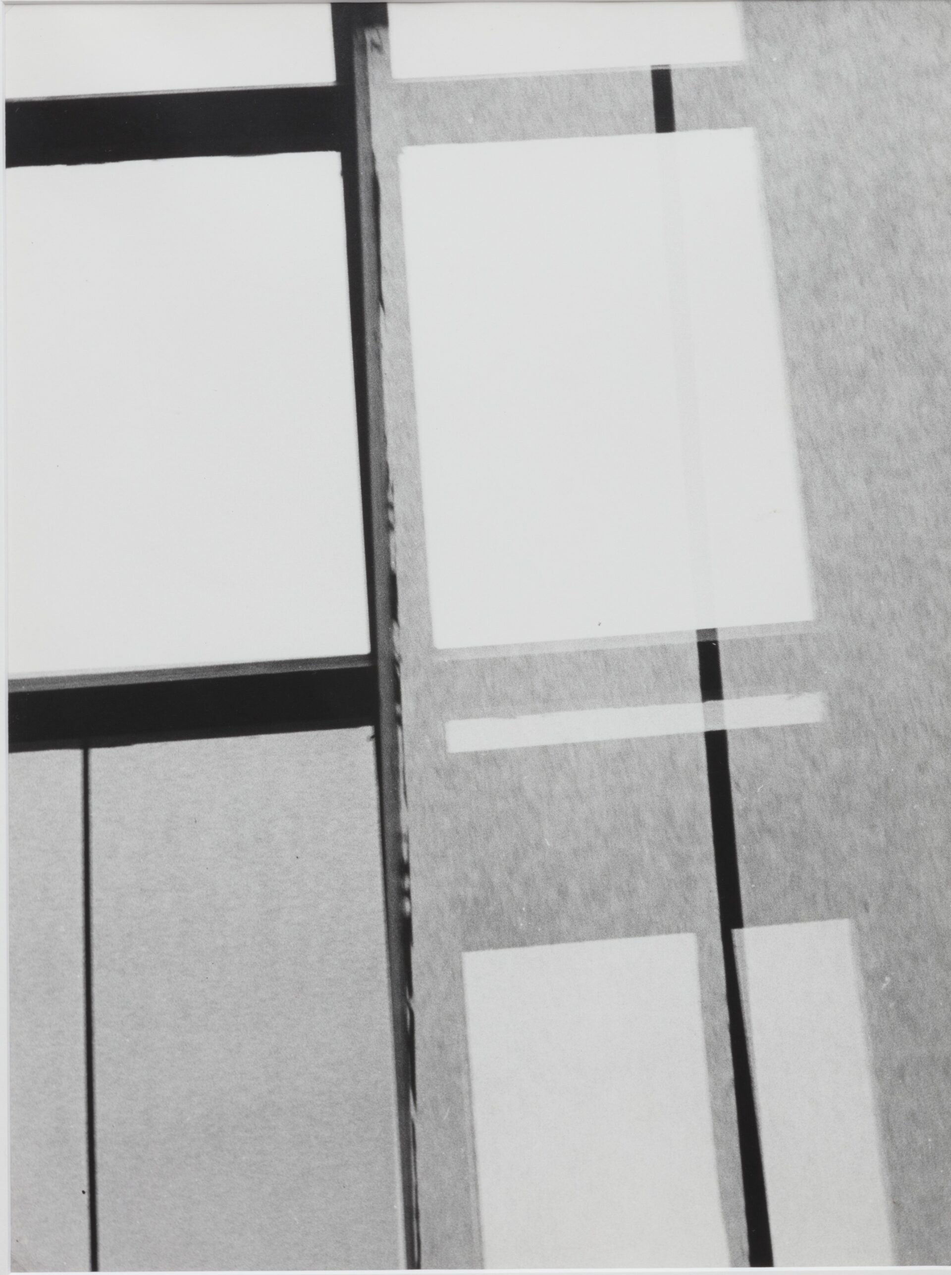Descrição da imagem: A fotografia vertical em preto e branco mostra uma composição geométrica e abstrata. Tem formas retangulares em diferentes tons de cinza. Na parte superior e inferior, há retângulos brancos que sobrepõem o fundo cinza de textura granulada. No centro, uma linha vertical preta e outra mais fina branca, dividem o espaço. Fim da descrição. 

