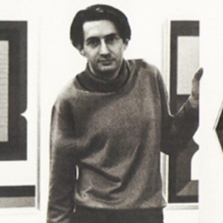 A imagem é um retrato em preto e branco de um homem jovem, de pele clara, em um ambiente de galeria ou exposição. O homem está posicionado no centro da imagem, ligeiramente inclinado para a direita, olhando diretamente para a câmera. Ele tem cabelo liso e escuro, penteado para o lado esquerdo da cabeça e de comprimento na altura das orelhas. Ele veste um suéter de gola rolê escuro (cinza ou preto) e calças escuras. Sua mão direita está levantada e apoiada na lateral de um quadro ou obra de arte que está fora do enquadramento, à direita. Ao fundo, há duas outras obras de arte penduradas em uma parede clara. Essas obras são abstratas e geométricas, apresentando formas retangulares escuras dentro de um fundo claro, com contornos bem definidos. A iluminação é uniforme.