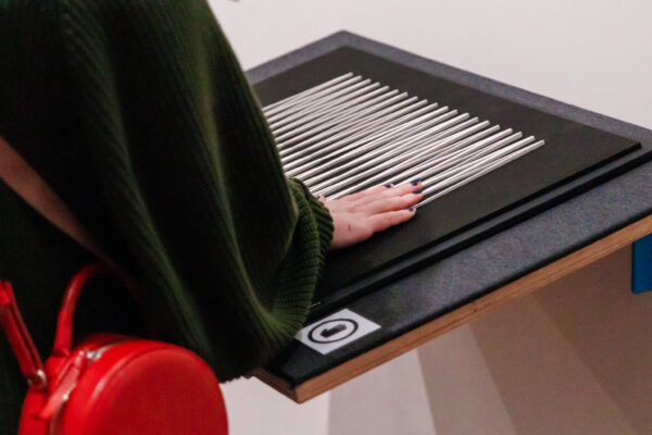 Descrição da imagem: a imagem é um close-up de uma pessoa interagindo com uma obra de arte ou objeto tátil em uma exposição. A mão da pessoa, com as unhas pintadas de azul marinho, está suavemente apoiada ou deslizando sobre uma superfície composta por várias hastes cilíndricas prateadas (que parecem ser de metal ou alumínio) alinhadas paralelamente sobre uma base escura (preta ou grafite). A obra está instalada sobre uma mesa de tampo preto, com a lateral de madeira clara. A pessoa veste uma malha ou suéter de tricô verde escuro e, no canto inferior esquerdo, é visível parte de uma bolsa pequena e redonda de cor vermelha vibrante. Um pequeno adesivo ou marcador branco com um ícone (possivelmente indicando uma área de interação) está fixado na lateral da mesa, perto da mão. Fim da descrição.