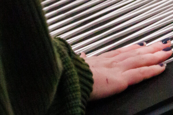 Descrição da imagem: a imagem é um close-up de uma pessoa interagindo com uma obra de arte ou objeto tátil em uma exposição. A mão da pessoa, com as unhas pintadas de azul marinho, está suavemente apoiada ou deslizando sobre uma superfície composta por várias hastes cilíndricas prateadas (que parecem ser de metal ou alumínio) alinhadas paralelamente sobre uma base escura (preta ou grafite). A obra está instalada sobre uma mesa de tampo preto, com a lateral de madeira clara. A pessoa veste uma malha ou suéter de tricô verde escuro e, no canto inferior esquerdo, é visível parte de uma bolsa pequena e redonda de cor vermelha vibrante. Um pequeno adesivo ou marcador branco com um ícone (possivelmente indicando uma área de interação) está fixado na lateral da mesa, perto da mão. Fim da descrição.