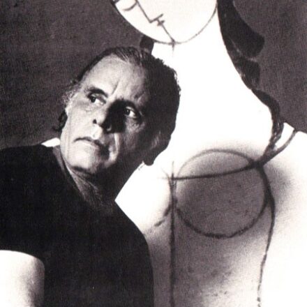 A imagem é um retrato em preto e branco de um homem de meia-idade, de pele clara, com uma obra de arte figurativa ao fundo. O homem está posicionado no lado esquerdo da imagem, virado para sua direita e olhando para fora do enquadramento, com uma expressão séria. Ele tem cabelo escuro, penteado para trás, e rugas visíveis na testa e ao redor dos olhos. Ele veste uma camiseta escura de gola redonda. À direita e atrás do homem, uma obra de arte bidimensional, de tamanho considerável, é visível. A obra apresenta uma figura feminina estilizada, predominantemente branca com contornos escuros. A figura tem um rosto angular, traços faciais simples e formas que sugerem o busto e os braços. O fundo geral é escuro e liso. A iluminação é dramática, vinda da frente e ligeiramente da esquerda, criando sombras profundas no rosto do homem e destacando a figura branca da obra de arte.