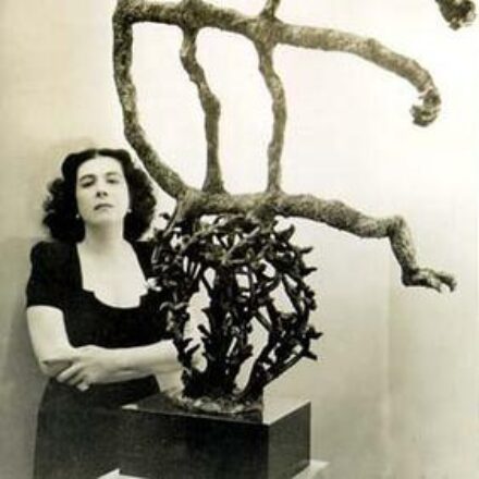 A imagem é uma fotografia em preto e branco de uma mulher e uma escultura abstrata. A mulher, de pele clara, está posicionada ao lado esquerdo da imagem, de pé, encostada em uma parede clara. Ela tem cabelo escuro, ondulado e volumoso na altura do ombro. Ela usa uma blusa escura de manga curta, com decote redondo. Seus braços estão cruzados na frente do corpo, e ela olha diretamente para a câmera com uma expressão séria. À direita da mulher, em primeiro plano, está uma escultura escura e abstrata. A escultura contém formas orgânicas e emaranhadas, que se assemelham a galhos, raízes ou espinhos entrelaçados. Da parte superior desta forma emaranhada, emerge uma estrutura que lembra uma figura humana estilizada ou um esqueleto. Essa estrutura tem "braços" e "pernas" finos e longos, feitos de material texturizado, que se estendem para os lados e para cima. O fundo é uma parede clara e lisa. A iluminação é suave.