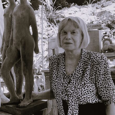 A imagem é um retrato em preto e branco de uma mulher idosa, de pele clara, em um ambiente externo que parece ser um jardim ou ateliê. A mulher está posicionada à direita do centro da imagem, olhando diretamente para a câmera com uma expressão neutra. Ela tem cabelo loiro claro, liso e curto, com franja. Ela veste uma blusa com um padrão estampado (animal print ou abstrato) e uma saia ou calça escura. À esquerda da mulher, e em primeiro plano, há duas esculturas de figuras humanas nuas, em tom escuro, de pé sobre pedestais. Elas parecem ser feitas de pedra. Outras esculturas e obras de arte, bem como vegetação e elementos de jardim, são visíveis no fundo desfocado, criando um ambiente repleto de arte e natureza. A iluminação é brilhante, sugerindo um dia ensolarado, e cria contrastes claros e escuros na imagem.