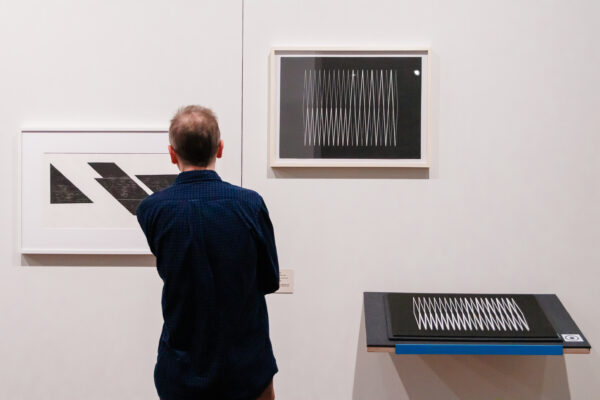 Descrição de imagem: a imagem mostra um visitante de costas em um espaço expositivo. O homem tem pele clara, é calvo no topo da cabeça, e está vestindo uma camisa social azul escura com mangas longas. Ele está observando duas obras de arte geométricas em preto e branco que estão penduradas em uma parede branca. A obra que ele está observando à esquerda apresenta faixas diagonais sólidas, enquanto a obra central exibe um padrão denso de linhas em zigue-zague brancas sobre fundo preto. Abaixo da obra de zigue-zague, sobre um pequeno pedestal, encontra-se uma representação tátil em relevo da mesma peça, indicando que a exposição é acessível. Fim da descrição.