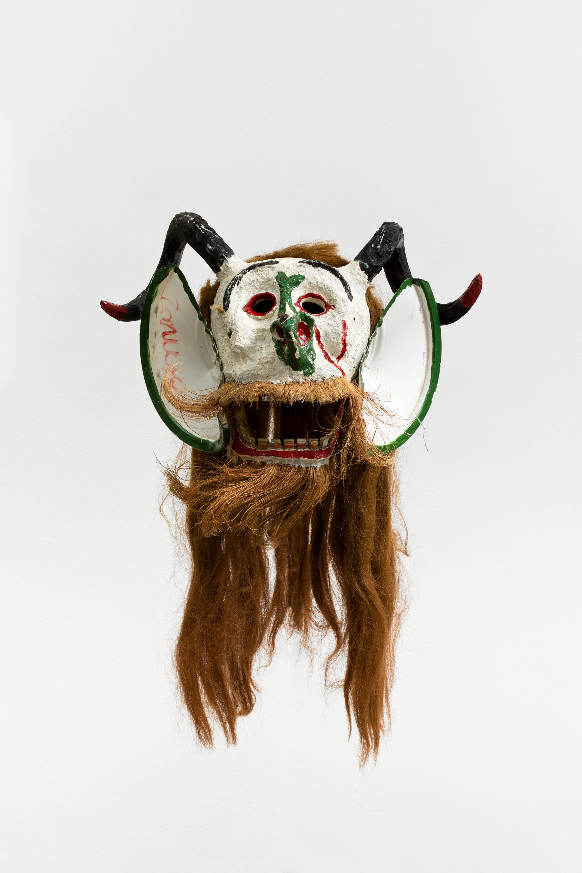 Descrição da imagem: Máscara tridimensional pintada. Vista de frente, a obra tem chifres pretos voltados para trás, curvados para baixo e as pontas vermelhas voltadas para cima. Atrás deles, há longos cabelos lisos e ruivos.
O rosto da máscara é branco, com sobrancelhas pretas, finas e arqueadas. Os olhos são vazados e contornados por um delineado vermelho. O nariz é verde e irregular. As orelhas, grandes e pontiagudas, são brancas, com uma linha verde nas bordas. Na orelha direita, em vermelho, está escrito “Zimar”. Na bochecha esquerda, duas linhas vermelhas contornam a maçã do rosto.
Os lábios são vermelhos e a boca, larga, está aberta, deixando à mostra dentes pequenos, quadrados, amarelados e avermelhados. O bigode e a barba lisos também são ruivos. Fim da descrição da imagem.
