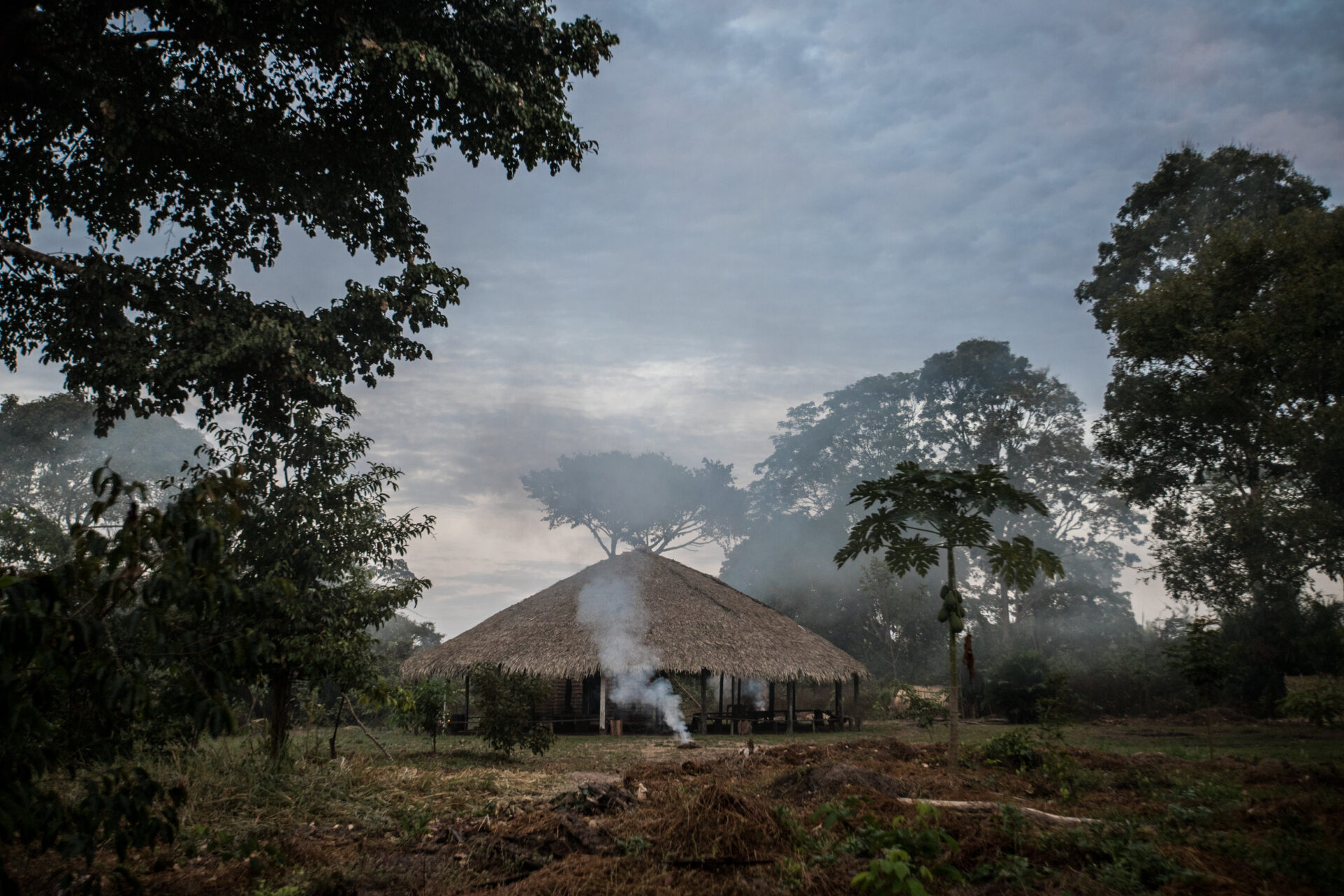 Descrição da imagem: Fotografia horizontal de uma construção em meio a uma floresta. Sob o céu azul, com nuvens brancas e em tons de cinza, em uma clareira há uma construção circular formada por colunas de madeira que sustentam um telhado de palha marrom em formato piramidal.
A estrutura está em uma área aberta, sobre um chão coberto por vegetação rasteira, capim, terra, folhas secas e galhos espalhados. Ao redor, árvores altas com copas frondosas e verdes.
À frente da construção, de um montinho de palha solta, sai uma fumaça branca que se eleva em direção ao céu. Fim da descrição da imagem.
