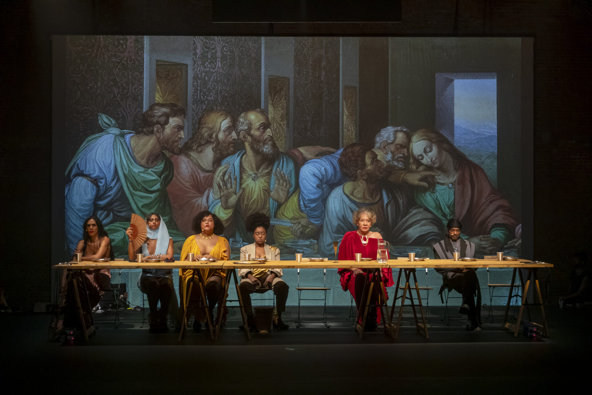 Descrição da imagem: Fotografia horizontal de quatro travestis, uma pessoa não-binária e um homem Cis, sentados atrás de uma longa mesa com tampo de madeira clara apoiado sobre cavaletes de madeira fina e escura. Sobre a mesa, há copos, pratos e uma jarra de água. Da esquerda para a direita: a primeira Travesti é negra de cabelos pretos, lisos e longos, usa roupa em tom rosado. A segunda Travesti é negra de sobrancelhas grossas, usa vestido azul-claro, com lenço na mesma tonalidade na cabeça, e segura um leque marrom aberto ao lado do rosto. A terceira Travesti é negra de cabelos castanhos, cacheados e volumosos, na altura dos ombros, usa roupa amarela com decote em V e colar de círculos amarelos. A pessoa não-binária é negra de cabelos castanhos, volumosos e presos em rabo de cavalo, usa blazer bege com detalhes dourados. Ao lado, há uma cadeira de madeira, com encosto vazado e pés metálicos finos. Ao lado dela, a última Travesti é branca de cabelos loiros e curtos, usa vestido vermelho com decote assimétrico, que deixa o ombro esquerdo à mostra e um colar dourado arredondado. Ao lado de mais uma cadeira vazia, está sentado o homem cis. Ele é preto e usa durag e blazer cinza com detalhes pretos. Ao lado dele, há outra cadeira. Ao fundo, há uma grande projeção de parte do quadro “A Última Ceia”, que ocupa toda a largura e altura da fotografia. Na projeção, há cinco homens e uma mulher, a maioria sentados à mesa. Da esquerda para a direita: um homem branco de cabelos castanhos, usa túnica azul com um tecido verde amarrado ao pescoço envolvendo o ombro esquerdo.

Um homem branco de cabelos loiros e longos, usa túnica vermelha com a gola dourada. Ele está com a mão direita apoiada no ombro de outro homem, que é branco, calvo, de cabelos e barba grisalhos. Usa túnica amarela e um manto azul sobre os ombros. Ele está com as mãos espalmadas para a frente.
O quarto homem também é branco, de cabelos e barba grisalhos. Usa roupa azul com tecido amarelo amarrado à cintura. Ele está parcialmente levantado, o dorso da mão direita sobre a cintura e o tronco encostado nas costas do último homem, também branco. Ele tem cabelos e barba castanho-escuros. Usa roupa azul.
A única mulher da cena é branca, tem longos cabelos loiros e usa túnica verde com tecido marrom sobre o ombro esquerdo. Ela está de olhos fechados e com a cabeça inclinada para a esquerda encostada na testa do homem que está parcialmente levantado. Os cinco homens estão com o olhar voltado para ela.
Na parede esquerda, ao fundo, faixas verticais com desenhos de arabescos rosados. À direita, uma passagem revela uma paisagem com montanhas e vegetação verdejante. Fim da descrição da imagem.
