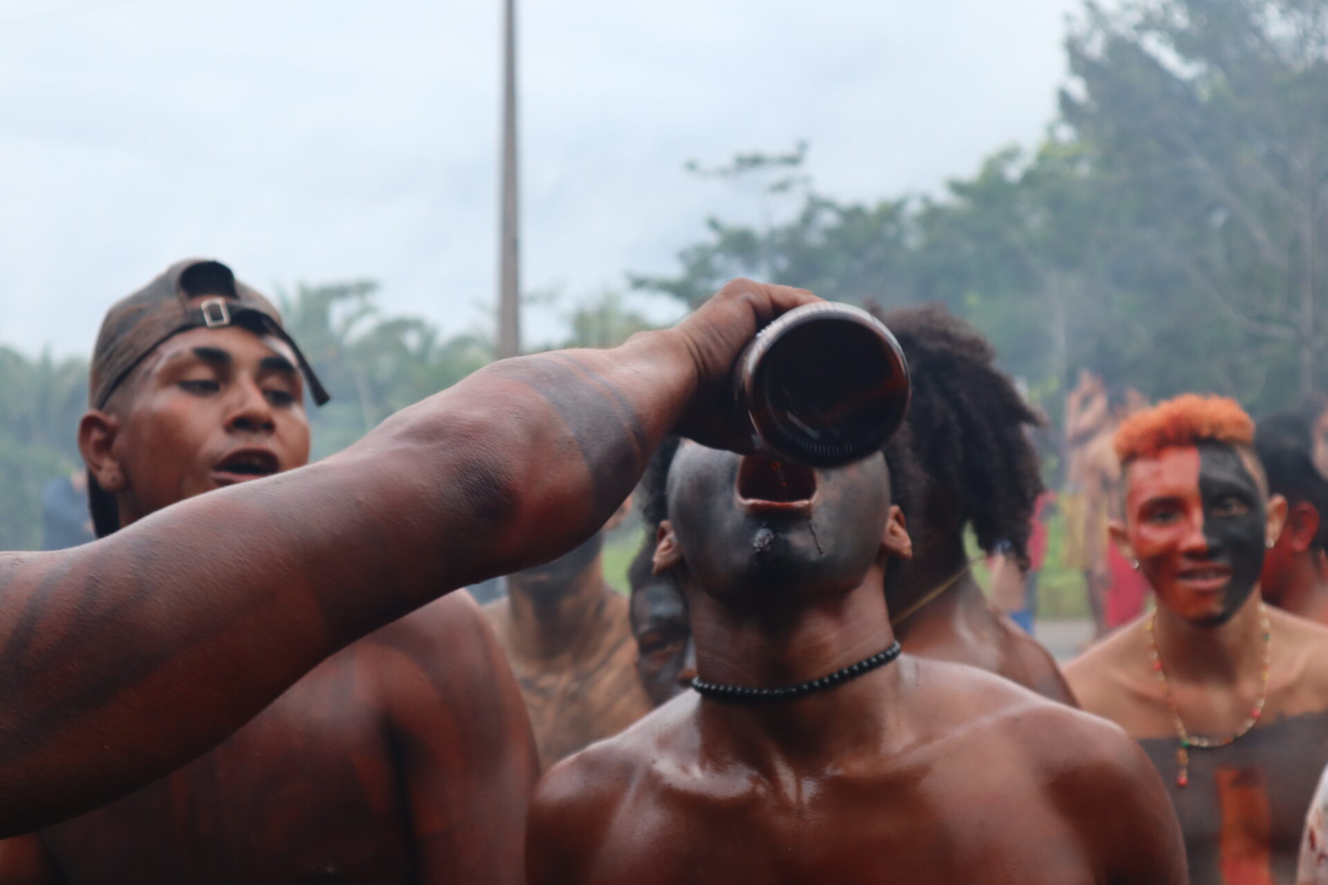 Descrição da imagem: Fotografia horizontal de um indígena tomando uma bebida. Ao centro, um homem de tronco nu, visto até o peito, está com o rosto pintado de preto e a cabeça inclinada para trás, com a boca aberta. À esquerda, uma pessoa, visível apenas pelo braço, despeja um líquido avermelhado de uma garrafa escura na boca do homem. O braço da pessoa está pintado de preto. À esquerda, atrás, outro homem de peito nu, com sobrancelhas grossas e curtas e boné marrom com a aba virada para trás, está com o olhar voltado para o homem ao centro. Ao fundo, à direita, um terceiro homem visto até o peito, com cabelos laranja; o rosto pintado de vermelho à direita e preto à esquerda, olhar fixo à frente e um leve sorriso. Ao fundo, desfocadas em meio a uma fumaça branca, há outras pessoas e, ao longe, árvores sob céu claro. Fim da descrição da imagem.
