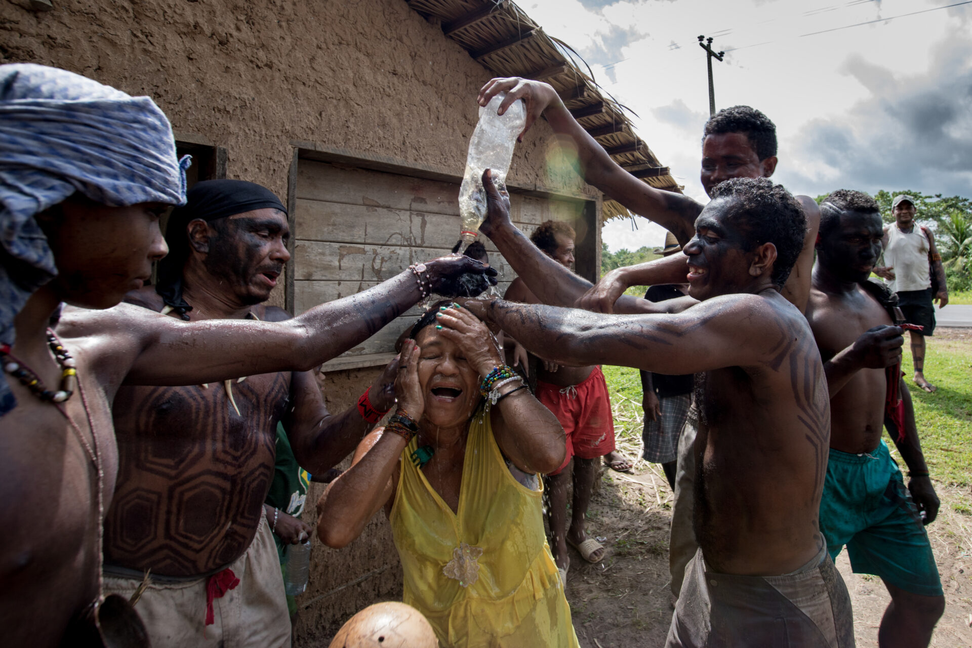 Descrição da imagem:
Fotografia horizontal de um grupo de indígenas. Ao centro, enquadrada da cintura para cima, há uma mulher com as mãos no rosto, olhos fechados e a boca aberta em um leve sorriso. Tem sobrancelhas um pouco ralas, nariz com a ponta arredondada e lábios médios. Usa regata amarela de alças um pouco largas e pulseiras coloridas nos dois punhos.
Homens estão ao redor dela, enquadrados da cintura para cima, segurando uma grande garrafa PET, da qual escorre água sobre a cabeça e o corpo da mulher.
À esquerda, um jovem com pano azul-claro na cabeça e peito nu, está com a mão esquerda perto da boca da garrafa. Ao lado, outro homem com pano preto na cabeça está com o tronco pintado com desenhos de hexágonos pretos.
À direita, o terceiro homem tem cabelos curtos, crespos e pretos. O tronco está nu, com pintura facial preta e pintura corporal em linhas angulares pretas. Usa bermuda cinza.
Atrás, o último homem, alto, é visto parcialmente; tem cabelos curtos, crespos e pretos, sobrancelhas grossas e segura a garrafa.
Ao fundo, uma casa revestida de barro com telhado de palha e alguns indígenas posicionados diante dela. O chão é de terra batida em tom terroso, intercalada com mato verde. O céu está acinzentado, com nuvens brancas.
Fim da descrição da imagem.
