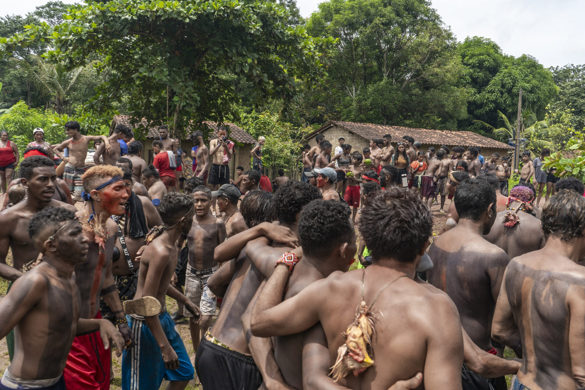 Descrição da imagem: Fotografia horizontal de um grande grupo de indígenas, reunidos sobre um chão terroso com trechos de grama verde. Muitos são homens, em pé, próximos uns dos outros, alguns vistos de costas e outros de frente. Parte do grupo está com os corpos pintados em preto e vermelho, usando bermudas vermelhas, azuis ou pretas, e adereços no pescoço, nos braços e na cabeça. Ao centro, quatro homens estão de costas, abraçados lateralmente; um deles usa um cordão fino com a cabeça de uma galinha nas costas. Ao fundo, duas construções de paredes beges e telhados marrons e muitas arvores com copas verdes. O céu está acinzentado. Fim da descrição da imagem.

