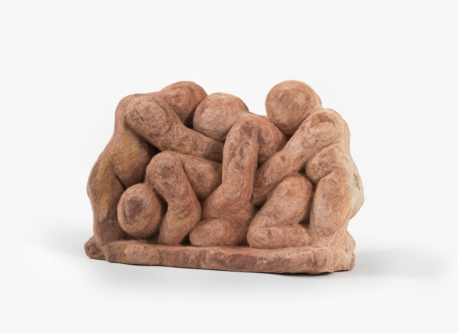 Descrição da imagem: Escultura composta por quatro figuras humanas esculpidas em arenito, uma rocha sedimentar. A peça tem coloração terracota, com variações entre o alaranjado e o marrom-claro. Da esquerda para a direita, uma figura em pé com o braço apoiado sobre outra figura ajoelhada, com o tronco e a cabeça inclinados para baixo. Atrás da figura ajoelhada, uma terceira figura está encostada e abraçada pela cintura da figura da frente. A quarta figura está posicionada atrás da terceira, também abraçada pela cintura da anterior. Estão esculpidas a partir de uma base plana com bordas irregulares. Fim da descrição da imagem.
