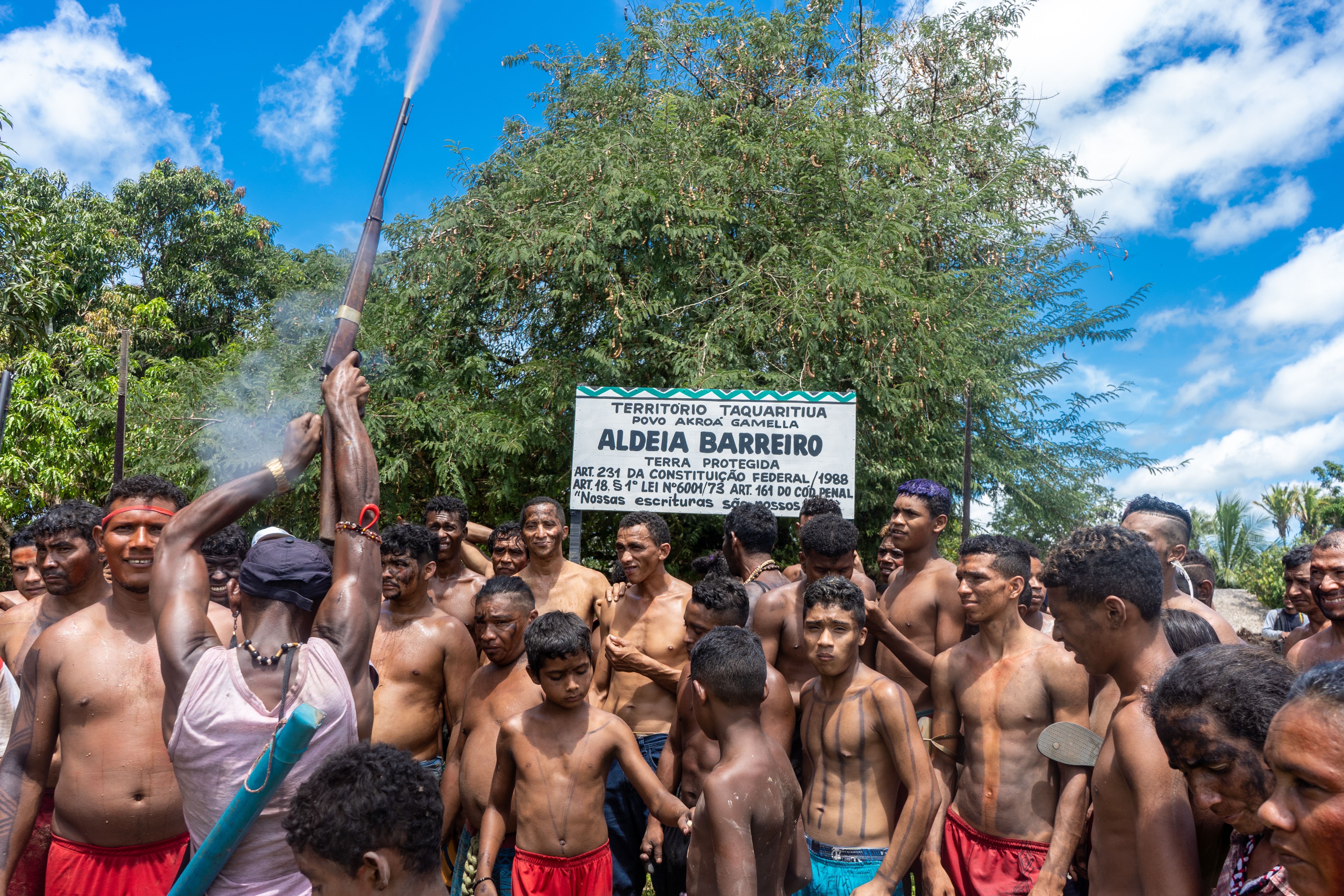 Descrição da imagem: Fotografia horizontal de um grupo de indígenas reunidos. Sob o céu azul com algumas nuvens brancas, a maioria está com o tronco nu e usa bermudas em diferentes cores, principalmente vermelhas e azuis. As pessoas estão de pé, lado a lado, agrupadas. À esquerda, um homem está de costas, com os braços torneados erguidos, segurando uma espingarda marrom apontada para o alto. Do cano, sai uma fumaça densa em linha reta para cima; do gatilho, uma fumaça mais suave. Ele usa boné azul, cordão de círculos pretos e camiseta rosa-claro. Está com um cilindro azul amarrado por uma corda que vai do ombro direito até a cintura.
Atrás do grupo, ao centro, uma placa branca com uma linha em zigue-zague verde no topo e, abaixo, letras grandes e azuis: Território Taquaritiua. Povo Akroá-Gamella. Aldeia Barreiro. Terra protegida. Artigo duzentos e trinta e um da Constituição Federal de mil novecentos e oitenta e oito. Artigo dezoito, parágrafo primeiro, Lei número seis mil e um de mil novecentos e setenta e três. Artigo cento e sessenta e um do Código Penal. Nossas escrituras são nossas. Ao fundo, há grandes árvores de copa esverdeada. Fim da descrição da imagem.
