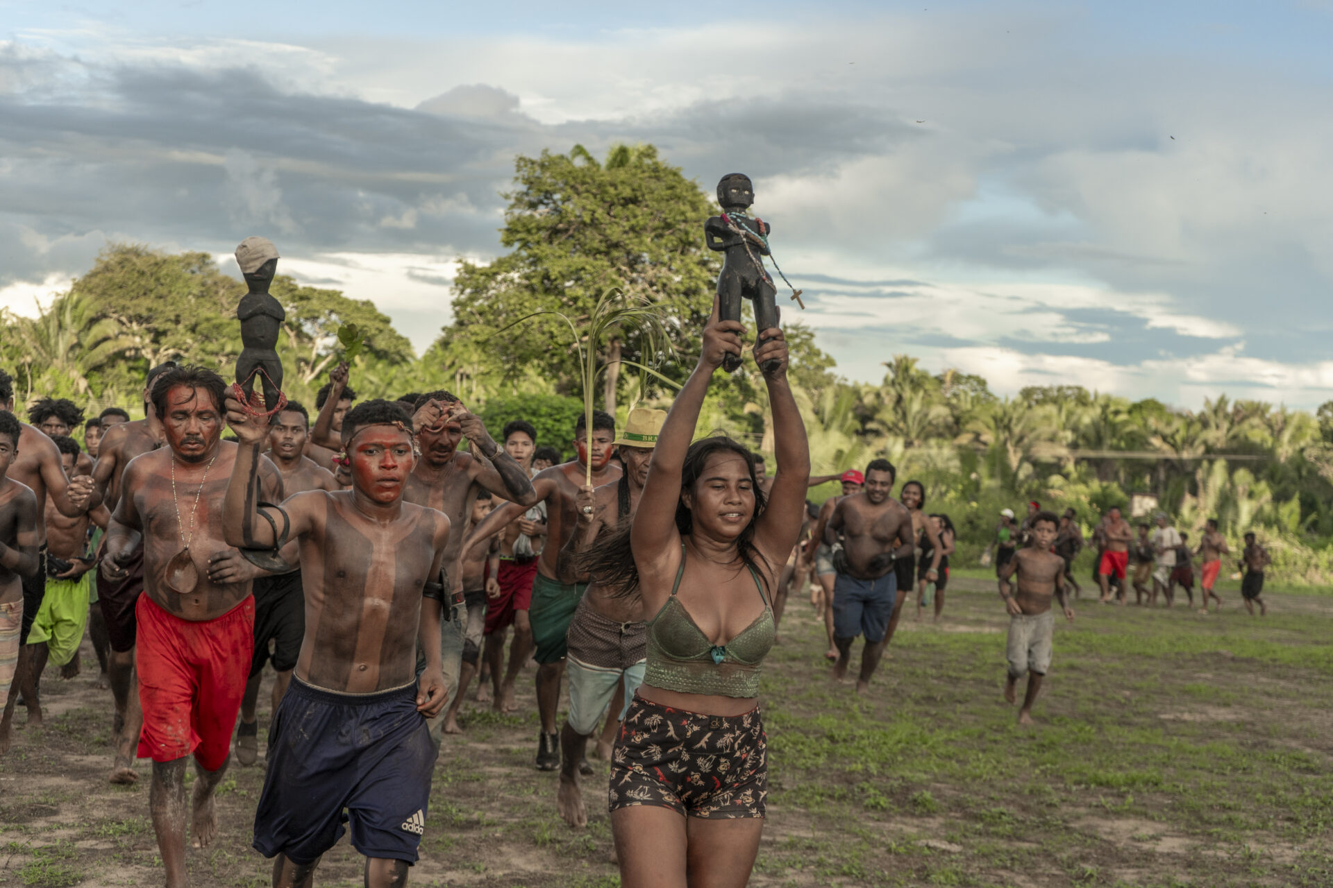 Descrição da imagem: Fotografia horizontal de um grupo de indígenas que avança à frente. Sob o céu azul com nuvens acinzentadas e largas, à frente do grupo, à esquerda, um menino está com o braço direito erguido, segurando um boneco nu, pintado de preto e com um turbante bege na cabeça. O menino tem cabelos curtos, lisos e pretos. Usa pintura facial vermelha e pintura corporal preta, com listras finas no braço e duas listras largas verticais que vão do peito até abaixo do abdômen. Usa bermuda azul. Ao lado, no centro, uma mulher está com os braços erguidos acima da cabeça, segurando pelas pernas outro boneco nu, pintado de preto. Ele está com os braços cruzados à frente e usa guias no pescoço. A mulher tem cabelos pretos, lisos e longos. Usa top verde e shorts estampado em preto e vermelho. A maioria das pessoas está com o tronco nu e o corpo coberto por pinturas em preto e vermelho. O solo é coberto por grama baixa, e ao fundo, há árvores de folhagens verdes. Fim da descrição da imagem.
