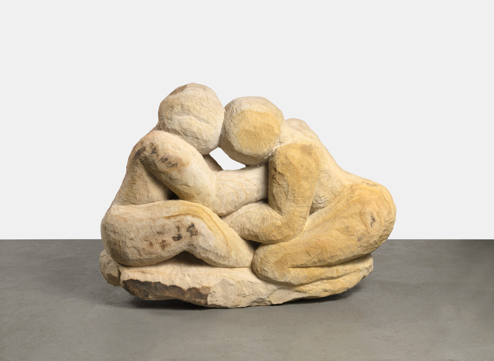 Descrição da imagem: Escultura composta por duas figuras humanas esculpidas em arenito, uma rocha sedimentar.
A peça tem coloração amarelada, com variações em marrom-claro e áreas mais escuras. De perfil e voltadas uma para a outra, a da esquerda está sentada e a da direita ajoelhada. Os troncos estão inclinados para frente, com as cabeças encostadas e os braços apoiados um no outro. Estão esculpidas a partir de uma base plana com bordas irregulares. Fim da descrição da imagem.
