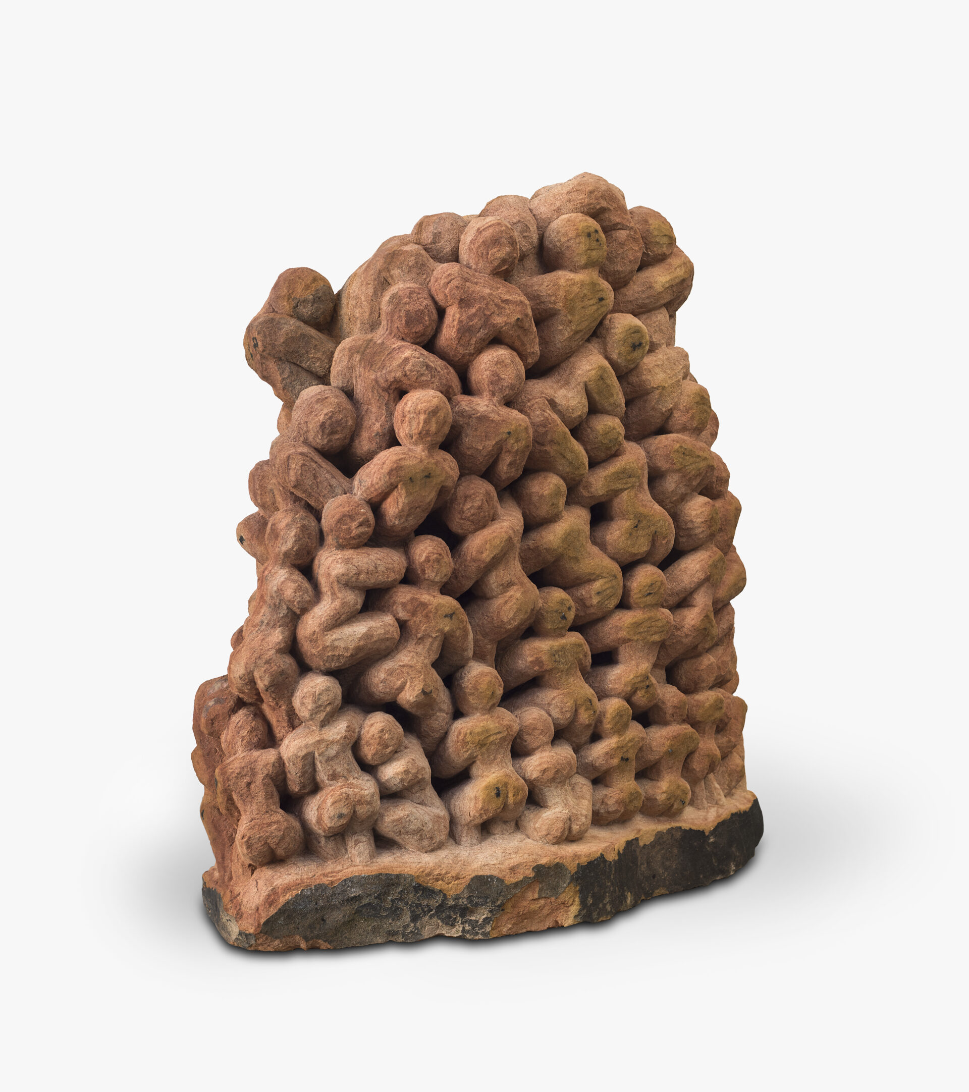 Descrição da imagem: Escultura vertical composta por figuras humanas aglomeradas esculpidas em arenito, uma rocha sedimentar. A peça tem cor terracota, com leves variações entre o alaranjado e o marrom-claro. As figuras têm corpo compacto, com cabeça arredondada e braços, tronco e pernas volumosos. Estão agrupadas lado a lado e empilhadas, em posições eretas e outras levemente curvadas, esculpidas a partir de uma base escurecida e irregular. Fim da descrição da imagem.
