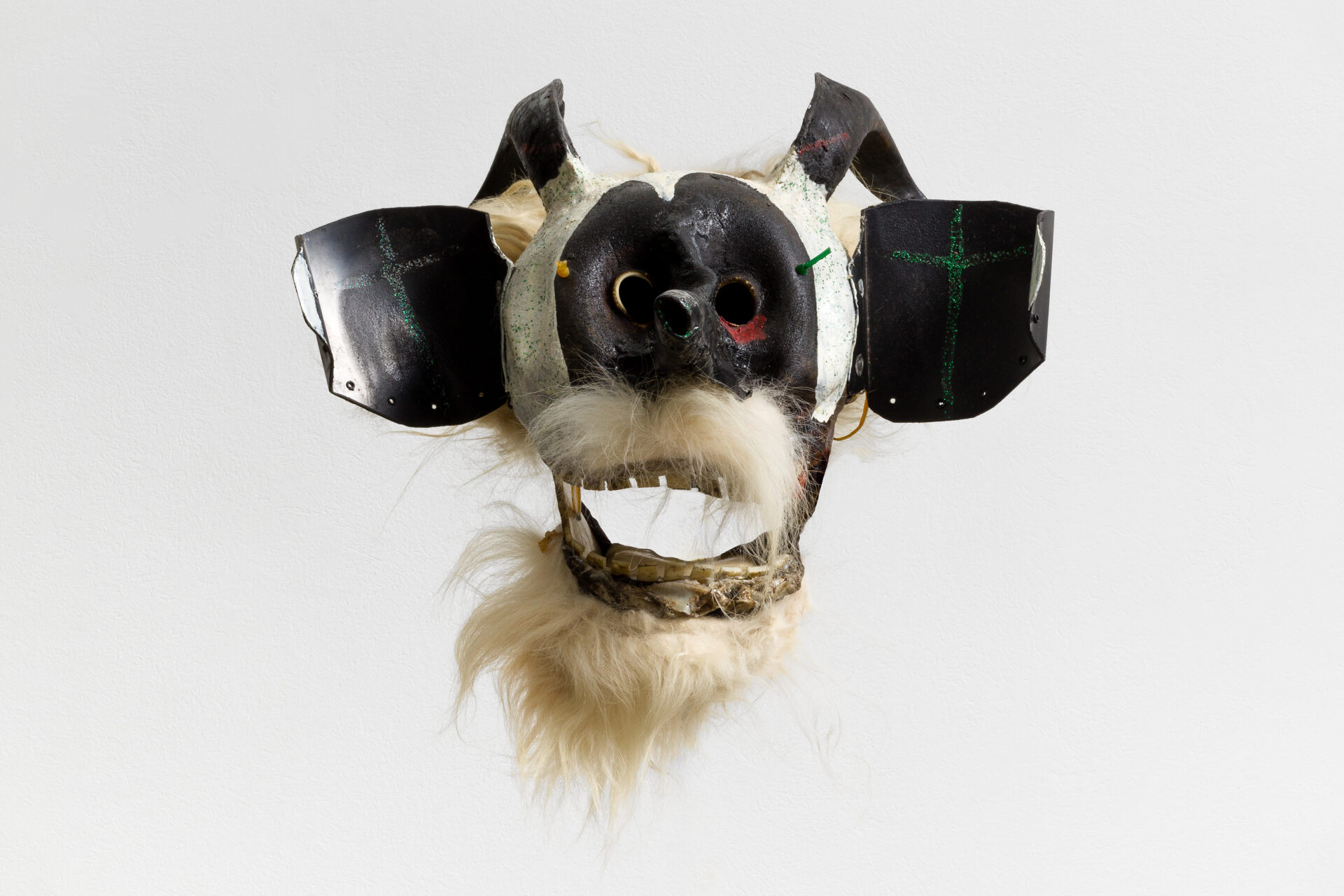 Descrição da imagem: Máscara tridimensional pintada. Vista de frente, a obra tem chifres pretos voltados para trás, curvados para baixo. Atrás deles, há cabelos brancos curtos, lisos, de textura macia, semelhante à pelúcia. O rosto é pintado de branco, com uma mancha arredondada preta ao redor dos olhos e do nariz. Os olhos são vazados: o esquerdo tem uma pequena mancha vermelha na pálpebra inferior; o direito, uma linha fina amarela ao redor. Na altura da têmpora esquerda, há um cordão verde inserido em um pequeno furo; na direita, um cordão amarelo. As orelhas são pretas e quadradas, com bordas irregulares, e em cada uma há uma cruz verde desenhada. A boca é larga e está aberta, deixando à mostra dentes quadrados, pequenos e amarelados. Tem bigode e barba brancos, cheios e lisos, de textura macia, semelhante à pelúcia. Fim da descrição da imagem.
