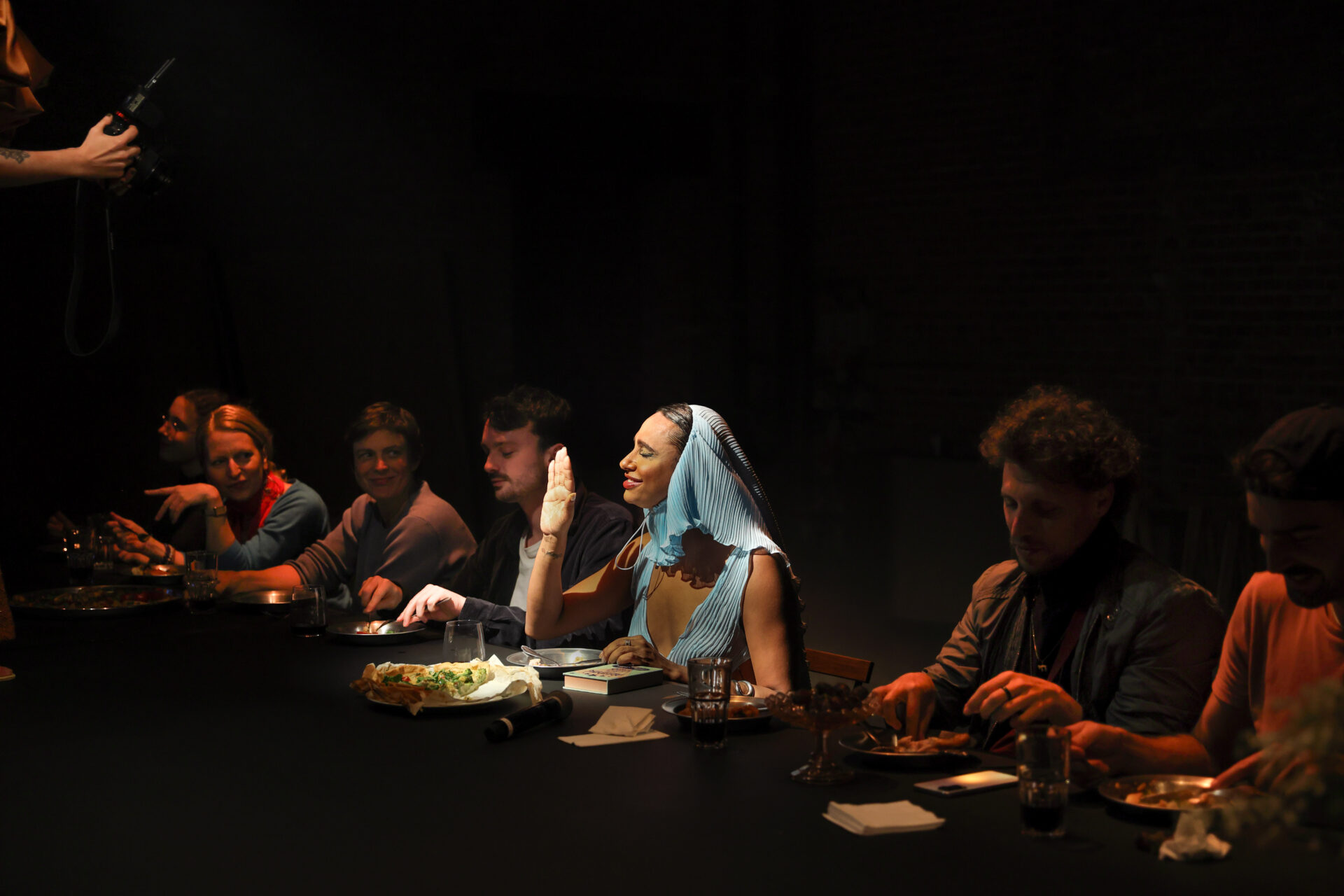 Descrição da imagem: Fotografia horizontal da Travesti negra de sobrancelhas grossas com o vestido e lenço azul-claro e algumas pessoas do público sentadas a uma mesa com tampo preto. Sobre a mesa, há um microfone, pratos, copos, talheres, alimentos, guardanapos e um livro. Da esquerda para a direita: Uma mulher branca de cabelos castanhos com o olhar voltado para a frente. Usa roupa preta. Uma mulher branca de cabelos lisos e loiros, com blusa azul de mangas longas e lenço vermelho ao redor do pescoço. Segura um talher com a mão direita, o cotovelo esquerdo apoiado sobre a mesa e o indicador erguido à frente. Uma mulher branca de cabelos castanhos e curtos, com camiseta cinza de mangas longas, está com um sorriso de lábios unidos. Um homem branco de cabelos curtos e bigode preto, com camiseta branca sob camisa cinza aberta, segura talheres e está com o olhar voltado para o prato à frente. A Travesti está com os olhos fechados e um sorriso, com a mão direita espalmada à frente e a esquerda apoiada sobre o livro. A mulher branca de cabelos lisos e loiros e a pessoa branca de cabelos castanhos e curtos, estão olhando para a Travesti. Um homem branco de cabelos castanhos curtos e cacheados, com jaqueta preta. Está com as mãos próximas ao prato e o olhar voltado para ele. Um homem branco de boné virado para trás e barba preta, com camisa cor de laranja, segura talheres e olha para o prato. No canto superior esquerdo, parte de um braço aparece com a mão segurando uma câmera fotográfica. O fundo está escuro. Fim da descrição da imagem.

