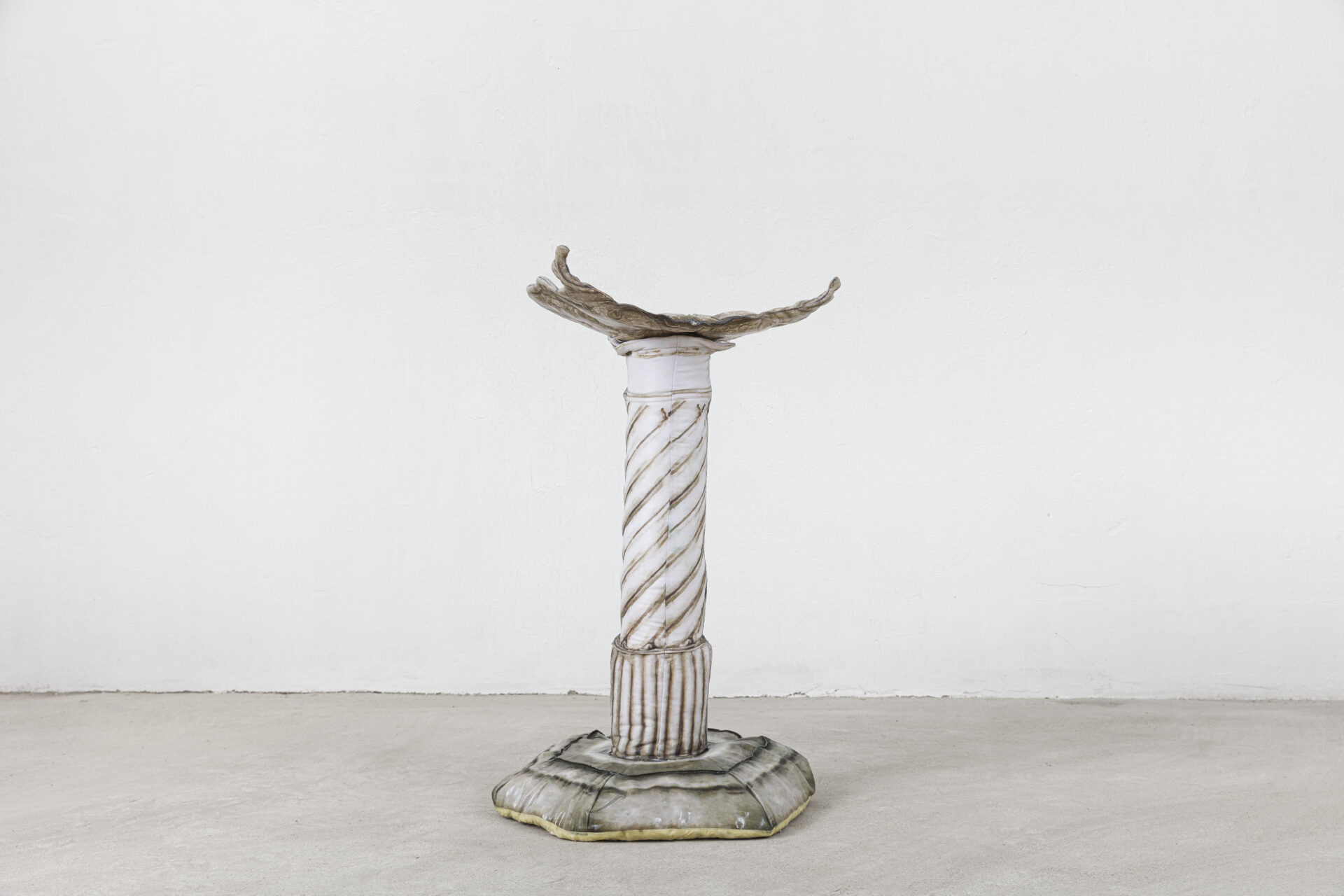 Descrição da imagem: Escultura de uma coluna vertical de jardim. Ela é composta por três partes: base, corpo e topo. O topo é uma peça branca, fina, irregular e horizontal, levemente arqueada para cima, com bordas assimétricas. O corpo central é uma coluna cilíndrica branca e alongada, com linhas espiraladas em marrom e linhas verticais na parte inferior. A base é larga e circular, de contorno irregular, acinzentada, com nuances esbranquiçadas e borda fina amarelada. Fim da descrição da imagem.
