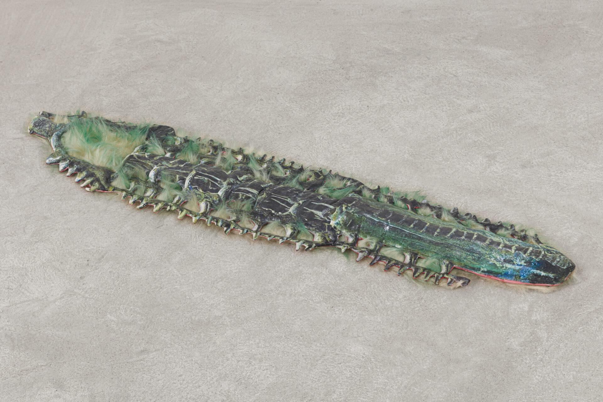 Descrição da imagem: Escultura plana e achatada feita de cimento e resina, em tons de verde-azulado. É rígida e levemente espessa. A forma geral se assemelha à parte dorsal estilizada de um inseto.
A peça é alongada e larga, com uma bifurcação na extremidade superior formada por duas pontas arredondadas. Abaixo dessa área, duas pontas maiores, também arredondadas. Nessa região, tufos de pelúcia tingida em verde e bege ocupam a superfície e contornam as bordas laterais de toda a peça.
A extremidade inferior é arredondada. Da área central até essa extremidade, a superfície é lisa e verde-azulada com poucas manchas pretas. No centro, uma linha clara percorre o trecho que vai da metade até a parte inferior da peça. Fim da descrição da imagem.
