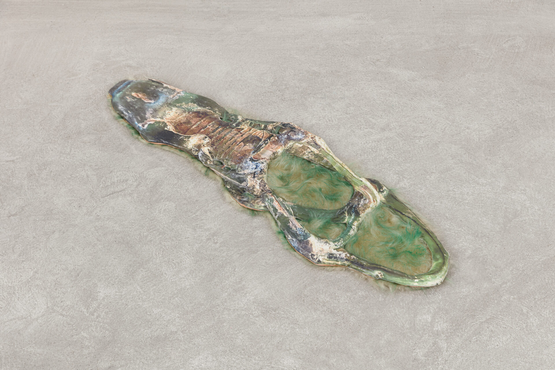 
Descrição da imagem: Escultura plana e achatada feita de cimento e resina, em tons de verde, bege, azul e preto. É rígida e levemente espessa. A forma geral se assemelha ao dorso alongado de um réptil estilizado.
A peça é alongada e ovalada, com formato irregular. A extremidade superior é afilada, com relevos irregulares manchados de verde, azul e preto. Linhas pretas em relevo estão dispostas horizontalmente no centro dessa área.
A extremidade inferior apresenta duas grandes cavidades arredondadas e irregulares na superfície, com interiores preenchidos por tufos de pelúcia tingida em verde e bege. A pelúcia também contorna partes das bordas laterais ao longo da peça. A superfície ao redor das cavidades apresenta variações de bege e verde. Fim da descrição da imagem.
