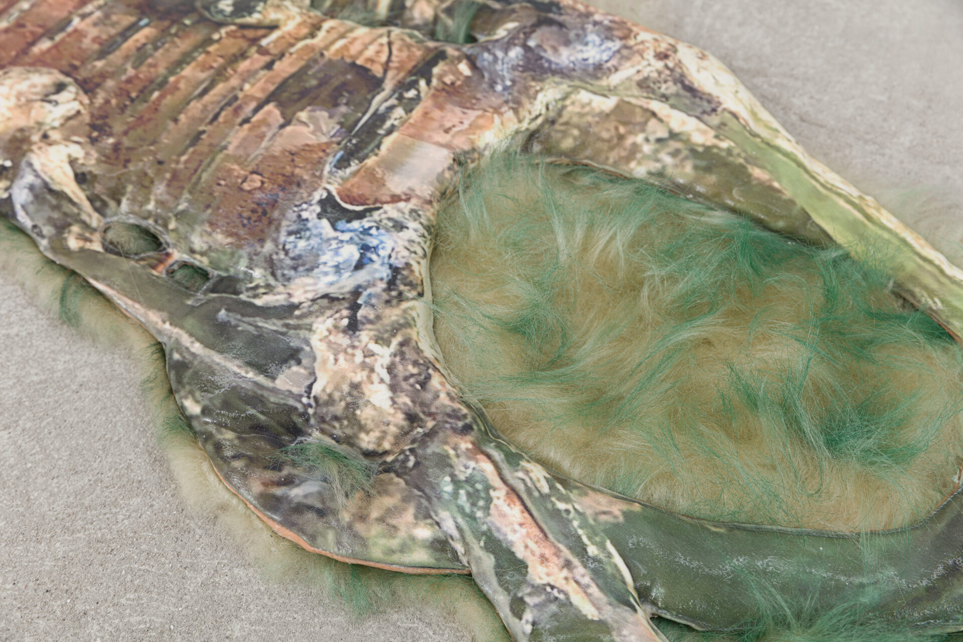 Descrição da imagem: Fotografia horizontal de uma área da escultura anterior, vista de perto. À esquerda, parte da superfície rígida com relevos irregulares manchados de verde, azul e preto. À direita, uma das cavidades arredondadas preenchida por tufos de pelúcia tingida em verde e bege. Fim da descrição da imagem.
