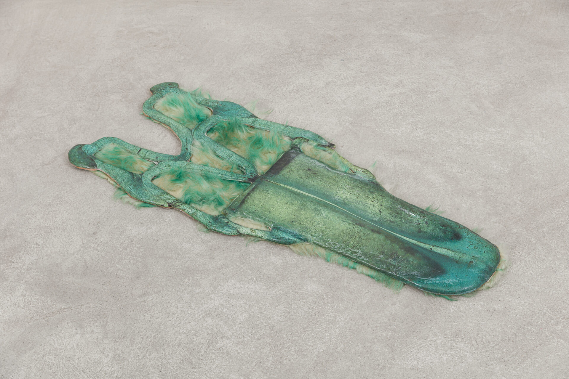 Descrição da imagem: Escultura plana e achatada feita de cimento e resina, em tons de verde e bege. É rígida e levemente espessa. A forma geral se assemelha ao dorso alongado de um réptil estilizado.
A peça é alongada e irregular, com bordas sinuosas formadas por pequenas saliências pontiagudas que se repetem ao longo de toda a lateral, assemelhando-se a dentes pontudos. Ao centro apresenta camadas e relevos irregulares em preto e cinza, intercalados por áreas em verde.
Na extremidade superior, tufos de pelúcia tingida em verde e bege ocupam parte da superfície. Na extremidade inferior, linhas horizontais em degradê de verde, azul e preto compõem a área final da peça. Fim da descrição da imagem.
