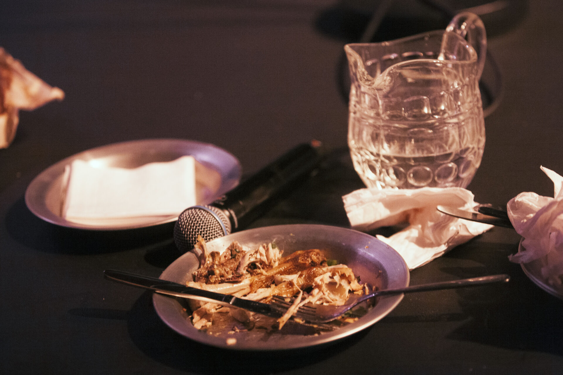 Descrição da imagem: Fotografia horizontal de uma parte da mesa preta.
Sobre a superfície, há dois pratos metálicos, um pequeno copo de vidro com formato de jarra, um microfone preto e guardanapos brancos. No prato ao centro, há restos de frango e ossos, com um garfo e uma faca dispostos sobre ele. O microfone está encostado à borda do prato. Atrás, o copo de vidro com água até a metade e, ao lado, um guardanapo amassado. À esquerda, o outro prato contém um pequeno monte de guardanapos dobrados. Fim da descrição da imagem.
