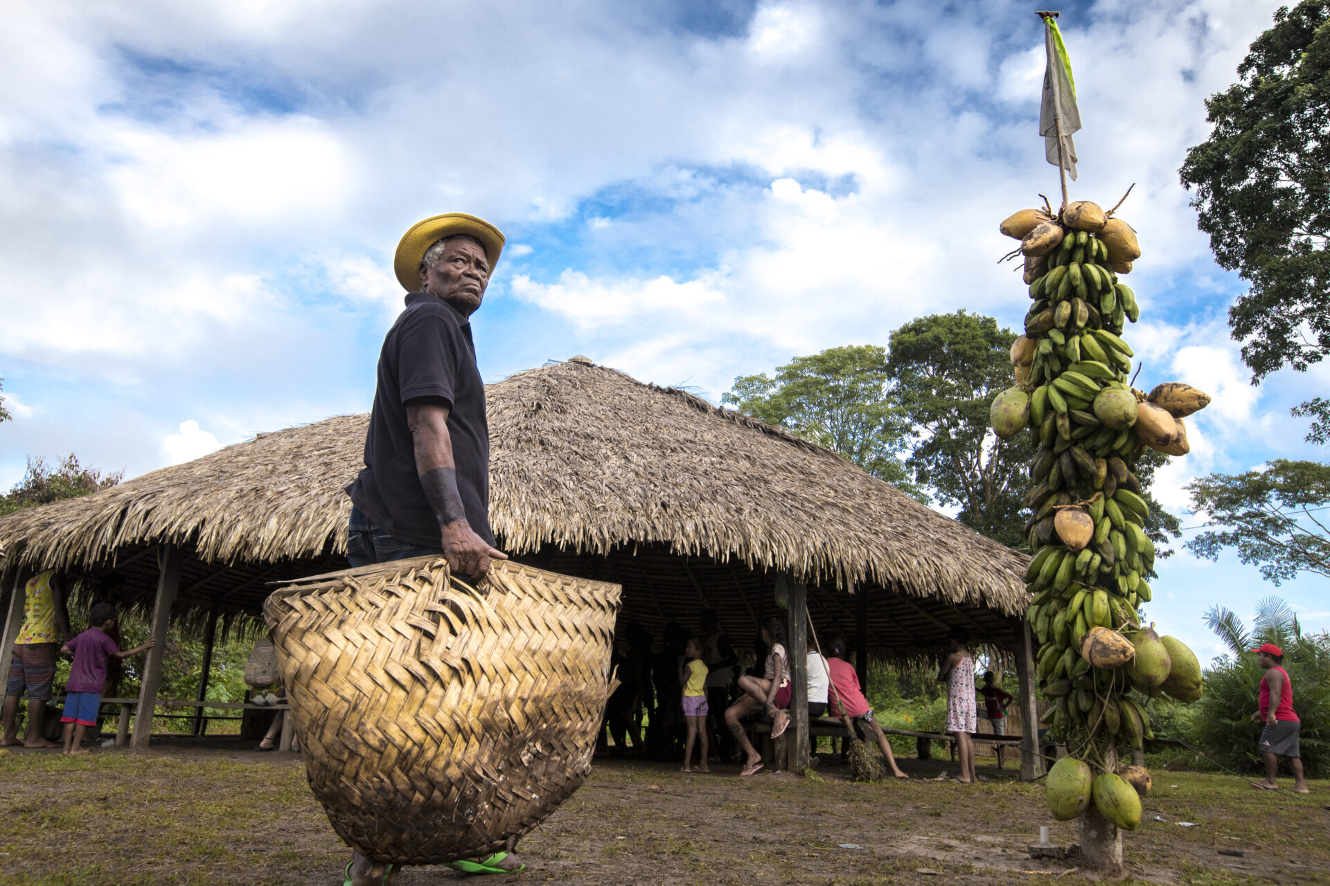 Descrição da imagem: Fotografia horizontal de um indígena perto da construção em meio à floresta. Sob o céu azul com nuvens brancas, à esquerda, visto parcialmente de costas, ele está em pé, com o tronco levemente virado para a direita e olhando por sobre o ombro. O homem tem cabelos grisalhos, olhos escuros e nariz largo. Usa chapéu amarelo, camiseta azul-escura, calça jeans e chinelos verdes. Segura com a mão direita uma grande cesta trançada de palha, de base arredondada.
À direita, bananas verdes, cocos e cacau estão presos a um tronco de árvore, com um bambu fino que sustenta uma bandeira branca e verde caída ao longo dele.
Ao fundo, a construção circular de colunas de madeira que sustentam o telhado piramidal de palha marrom, com muitas pessoas, algumas em pé e outras sentadas em bancos de madeira. O chão é de terra escura, com capim baixo e verde. Ao fundo, árvores altas com copas frondosas e verdes. Fim da descrição da imagem.
