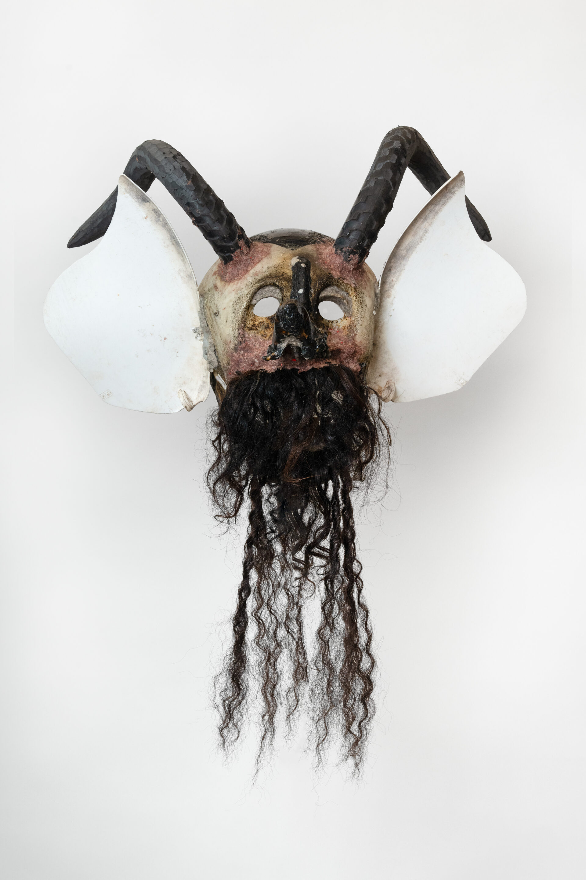 Descrição da imagem: Máscara tridimensional pintada. Vista de frente, a obra tem chifres pretos e compridos voltados para trás e curvados para baixo. Na base dos chifres, há uma mancha cor-de-rosa arredondada. A máscara é branca, com olhos vazados e nariz irregular preto. Nas bochechas, manchas cor-de-rosa. As orelhas são grandes e brancas. A boca está aberta, e dela saem cabelos cacheados castanho-escuros, que cobrem os dentes e a mandíbula, com algumas mechas se alongando para baixo. Fim da descrição da imagem.
