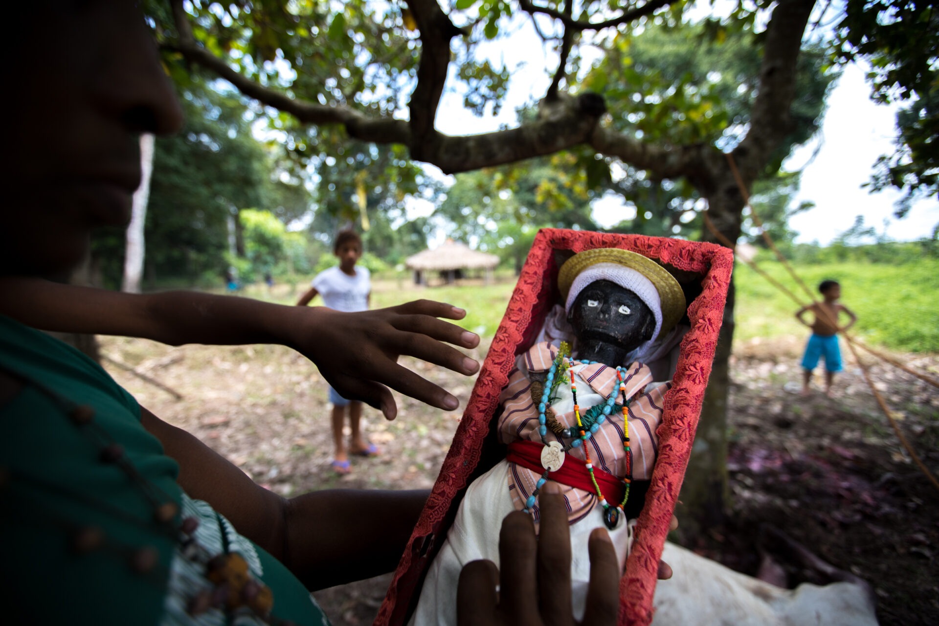 Descrição da imagem: Fotografia horizontal de um indígena segurando uma caixa com uma boneca. À esquerda, visto parcialmente de perfil direito, o indígena tem nariz alongado e lábios grossos, usa camiseta verde. Segura a caixa com a mão esquerda e, com a direita, toca a saia da boneca com os dedos indicador e médio.
A boneca é vista de perto dentro da caixa retangular coberta por uma renda vermelha. Tem pele preta e olhos brancos. Usa camisa rosa-claro com listras finas brancas e roxas, saia branca e faixa vermelha na cintura. Na cabeça, um chapéu dourado sobre um tecido branco. No pescoço, colares de contas, sendo uma azul-clara e outra com contas amarelas, pretas, vermelhas e verdes.
Atrás do indígena, o braço fino e esquerdo de uma pessoa aparece com a mão perto da caixa. Ao fundo, em desfoque, há mato baixo com árvores de copa verde, duas crianças indígenas e, ao longe, a construção circular com teto de palha sob o céu acinzentado. Fim da descrição da imagem.

