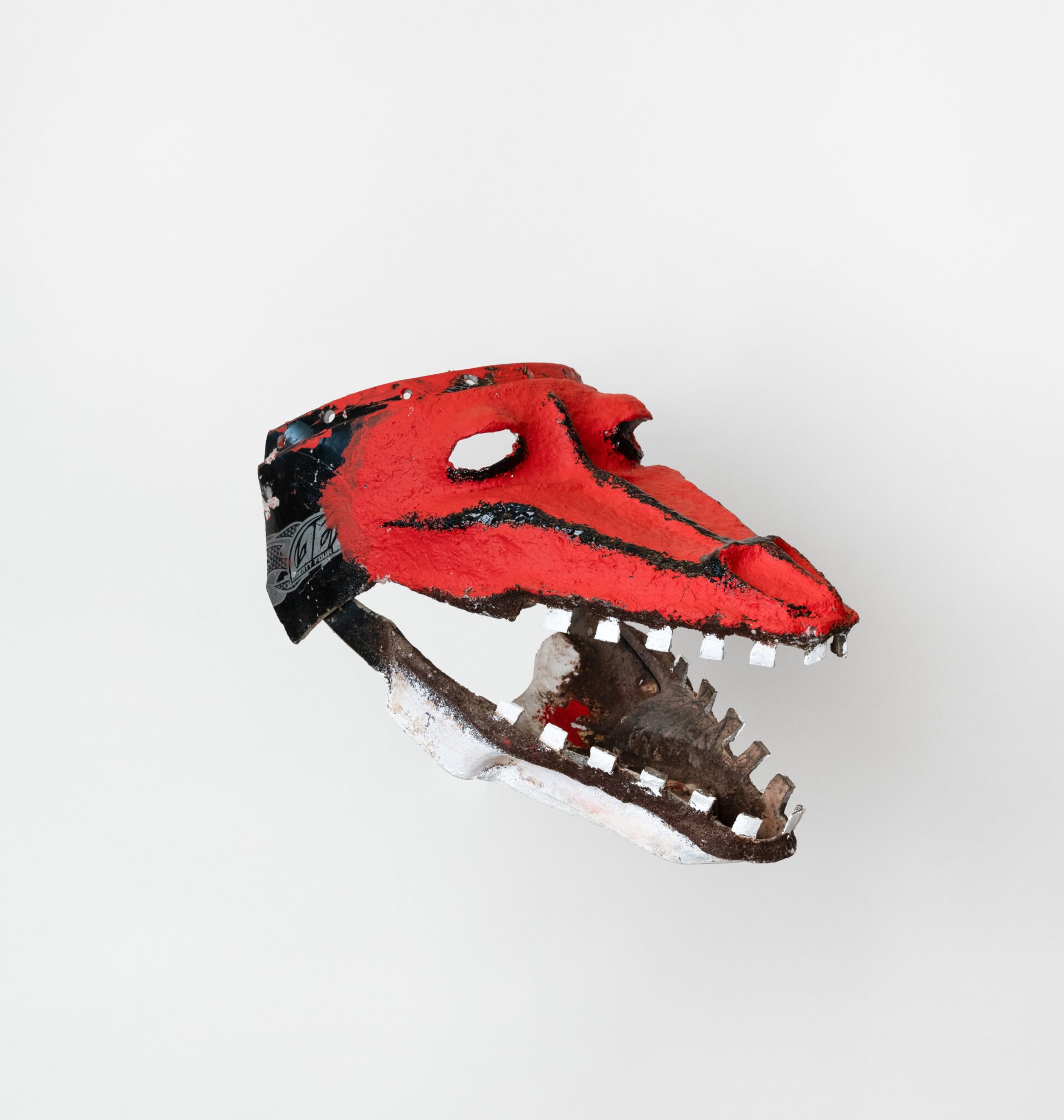 Descrição da imagem: Máscara tridimensional pintada. Vista de perfil para a direita, a obra tem a cara alongada na horizontal, que se prolonga em um focinho comprido. A máscara é vermelha, com olhos vazados e delineados em preto na parte inferior.    Uma linha preta parte da região entre os olhos e outra da área abaixo deles, ambas se estendendo até a extremidade do focinho, onde ficam narinas ovaladas. A boca está aberta, revelando interior marrom. Os dentes são brancos, pequenos, quadrados e espaçados, e a mandíbula é branca. Fim da descrição da imagem.

