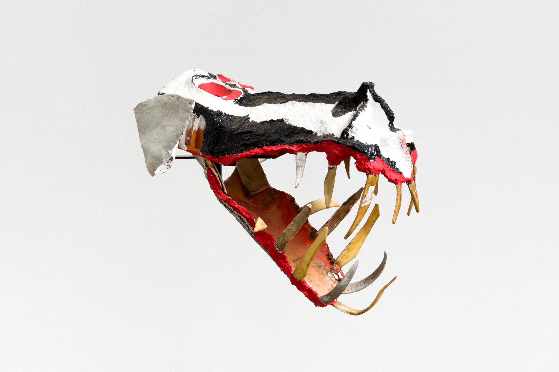 Descrição da imagem: Máscara tridimensional pintada. Vista de perfil para a direita, a obra tem a cara alongada na horizontal, que se prolonga em um focinho comprido.
Os olhos são arredondados e vermelhos, e as orelhas, triangulares em cinza.
A cara e o focinho são listrados de preto e branco, com uma pequena elevação próxima à extremidade do focinho.
A boca é contornada de vermelho e está aberta, deixando à mostra o interior alaranjado e os dentes prateados e dourados, finos e compridos. Fim da descrição da imagem.