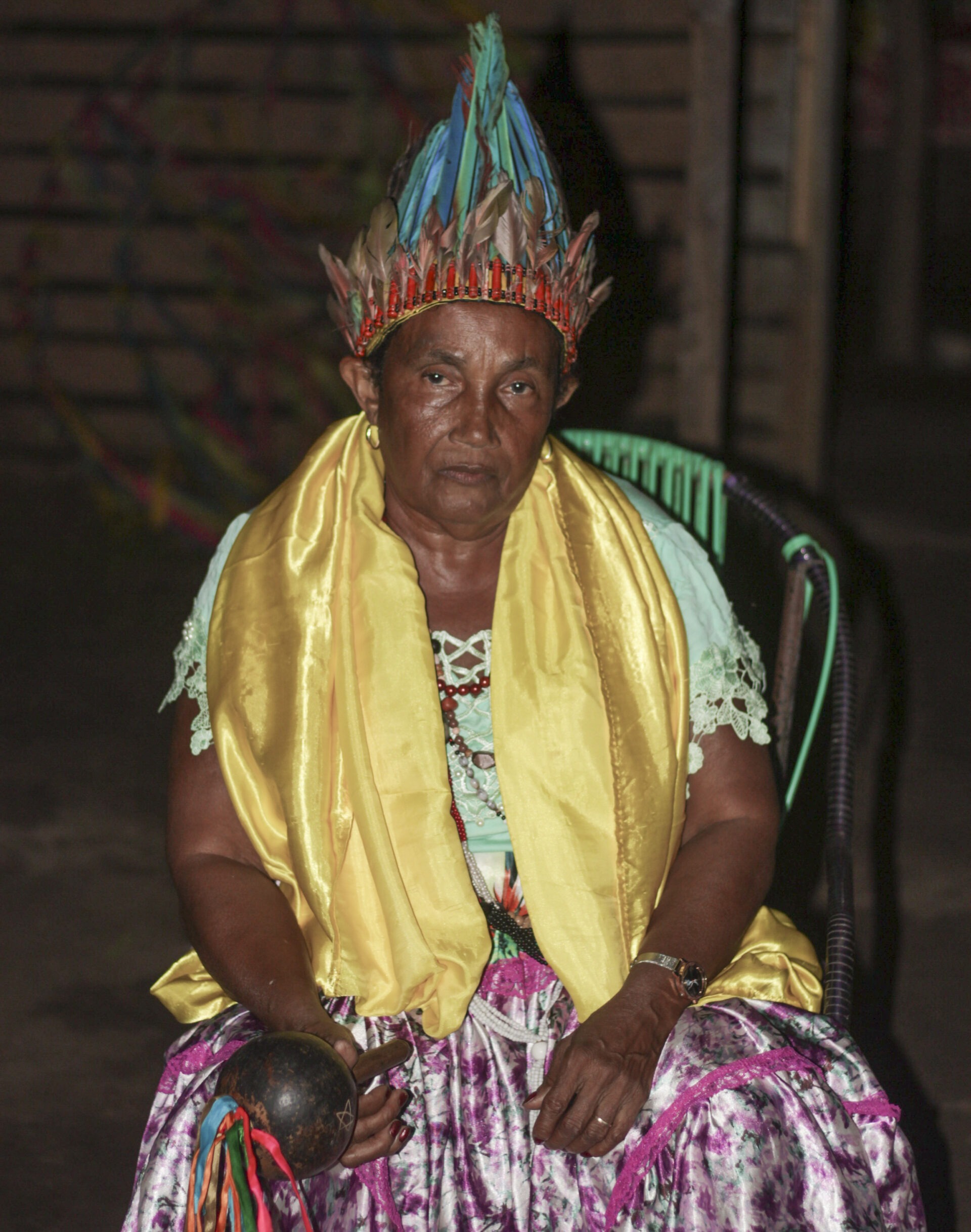 Descrição da imagem: Fotografia vertical da Pajé Craa Cwyj. Ao centro, enquadrada dos joelhos para cima, vista de frente, ela está sentada em uma cadeira metálica com encosto de fios plásticos verdes e roxos. Tem sobrancelhas grossas e curtas, olhos escuros e pequenos, nariz de ponta arredondada e lábios médios. Está com blusa verde-clara com rendas nas mangas curtas e uma saia floral em tons de rosa e verde.
Na cabeça, um cocar com penas em azul, vermelho e bege, presas a uma base bege com detalhes em vermelho; as penas se juntam e formam um detalhe afilado na ponta.
Sobre os ombros, um tecido amarelo cintilante. No punho esquerdo, usa um relógio de pulseira prateada e, no dedo anelar, uma aliança fina.
Na mão direita, segura um maracá marrom-escuro, com fitas nas cores vermelho, azul, verde e laranja presas ao cabo. Ao fundo, em desfoque, há uma parede de madeira. Fim da descrição da imagem.

