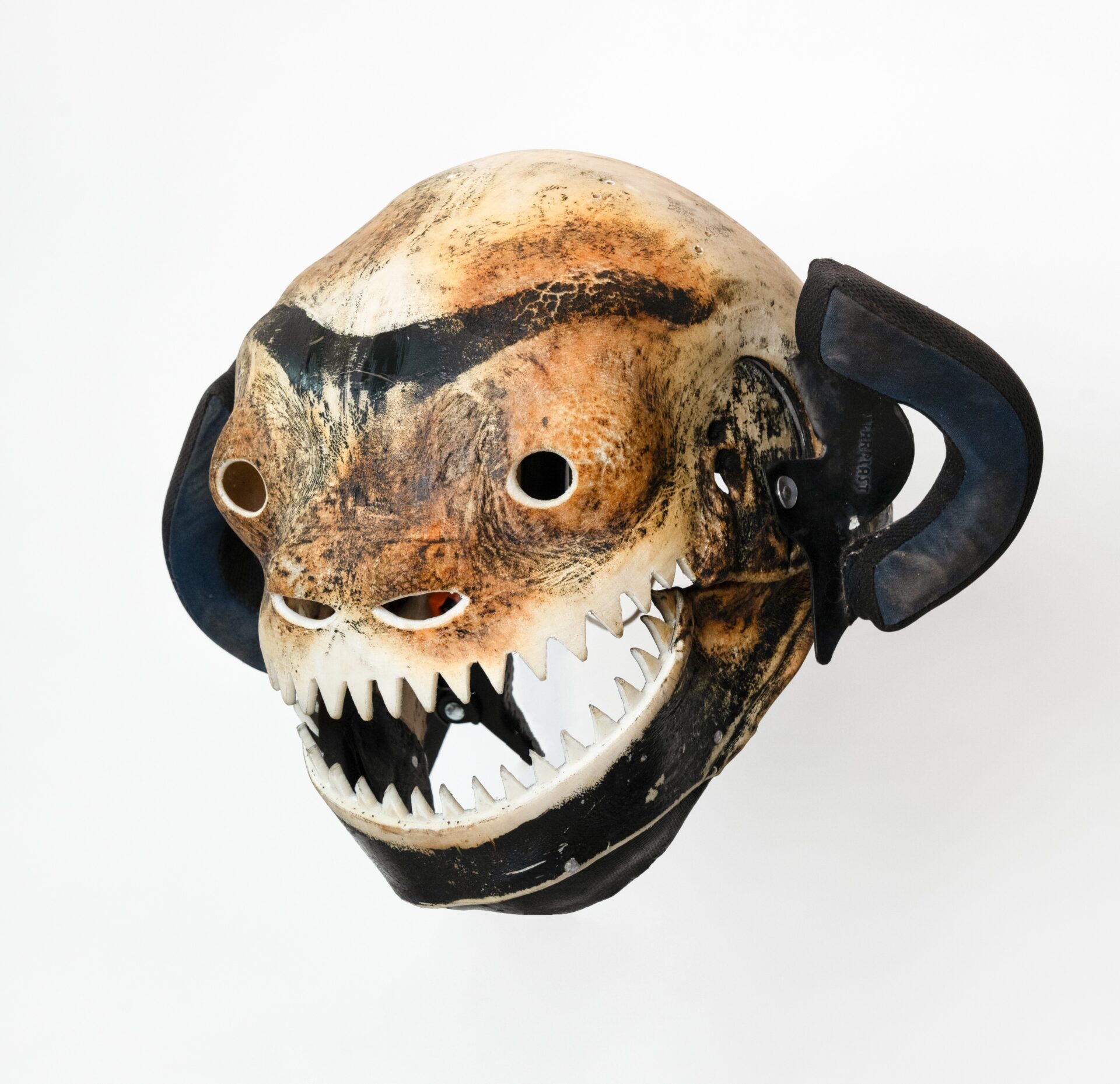Descrição da imagem: Máscara tridimensional pintada. Vista levemente de perfil para a esquerda, a obra tem estrutura arredondada em tons terrosos. As orelhas são formadas por pequenas estruturas em arco, acolchoadas em preto e azul-escuro. As sobrancelhas são unidas em uma longa linha preta, levemente arqueada. Os olhos são pequenos, redondos e vazados. As narinas consistem em duas formas ovais horizontais, pequenas e vazadas. A boca é larga e está aberta, com dentes brancos e triangulares. A mandíbula é preta. Fim da descrição da imagem.
