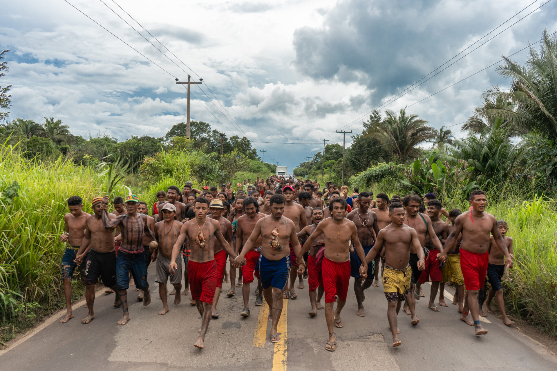 Descrição da imagem: Fotografia horizontal de um grande grupo de indígenas. Sob o céu azul com nuvens brancas, a maioria são homens que caminham lado a lado, de mãos dadas, organizados em fileiras que se estendem até o fundo da imagem. O grupo ocupa toda a largura de uma estrada asfaltada, marcada ao centro por duas linhas amarelas.
Os homens estão com o peito nu, alguns com pinturas de linhas grossas na vertical, do peito até abaixo do abdômen, e usam bermudas em diferentes cores: vermelhas, amarelas, azuis, cinza e jeans.
Alguns usam chapéus ou bonés; outros estão descalços ou de chinelos. Algumas mulheres estão no grupo, usando roupas coloridas. Uma está enquadrada do busto para cima e usa regata vermelha; outra usa cropped azul de alças finas, com detalhes cor-de-rosa e shorts jeans curtos.
À frente do grupo, quatro homens caminham de mãos dadas. Dois deles, à esquerda, usam cordões nos quais há a cabeça de galinhas amarradas.
A estrada é ladeada por vegetação alta e densa, com árvores e palmeiras. Postes com fios de energia estão alinhados ao longo da via. Fim da descrição da imagem.


