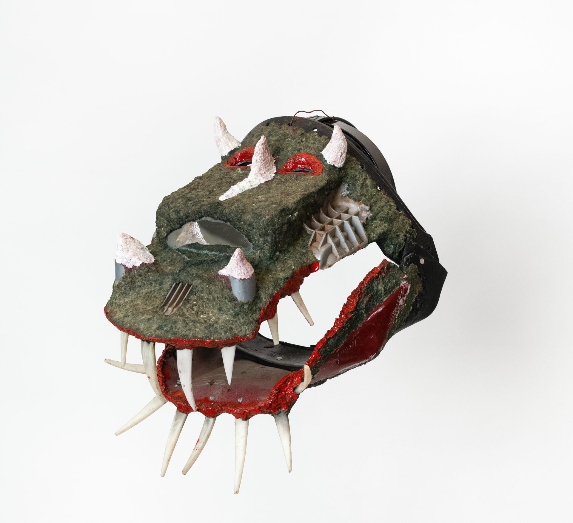 Descrição da imagem:  Máscara tridimensional pintada, que se assemelha à cabeça de um crocodilo. Vista levemente de perfil para a esquerdo, a obra tem a cara alongada na horizontal, que se prolonga em um focinho comprido. A máscara é pintada de verde, com textura irregular marcada por pequenas elevações, e tem olhos vermelhos. Acima deles, nas laterais, há dois pequenos chifres brancos e, entre os olhos, um terceiro chifre branco, do qual parte uma estreita mancha branca que desce pelo centro do focinho. O nariz é largo e quadrado, com uma narina semicircular vazada. À frente do nariz, nas laterais do focinho, há dois pequenos chifres brancos. O contorno da boca é vermelho, e ela está aberta, deixando à mostra dentes brancos, alongados e pontudos. Os dentes inferiores são inclinados para baixo. Fim da descrição da imagem.
