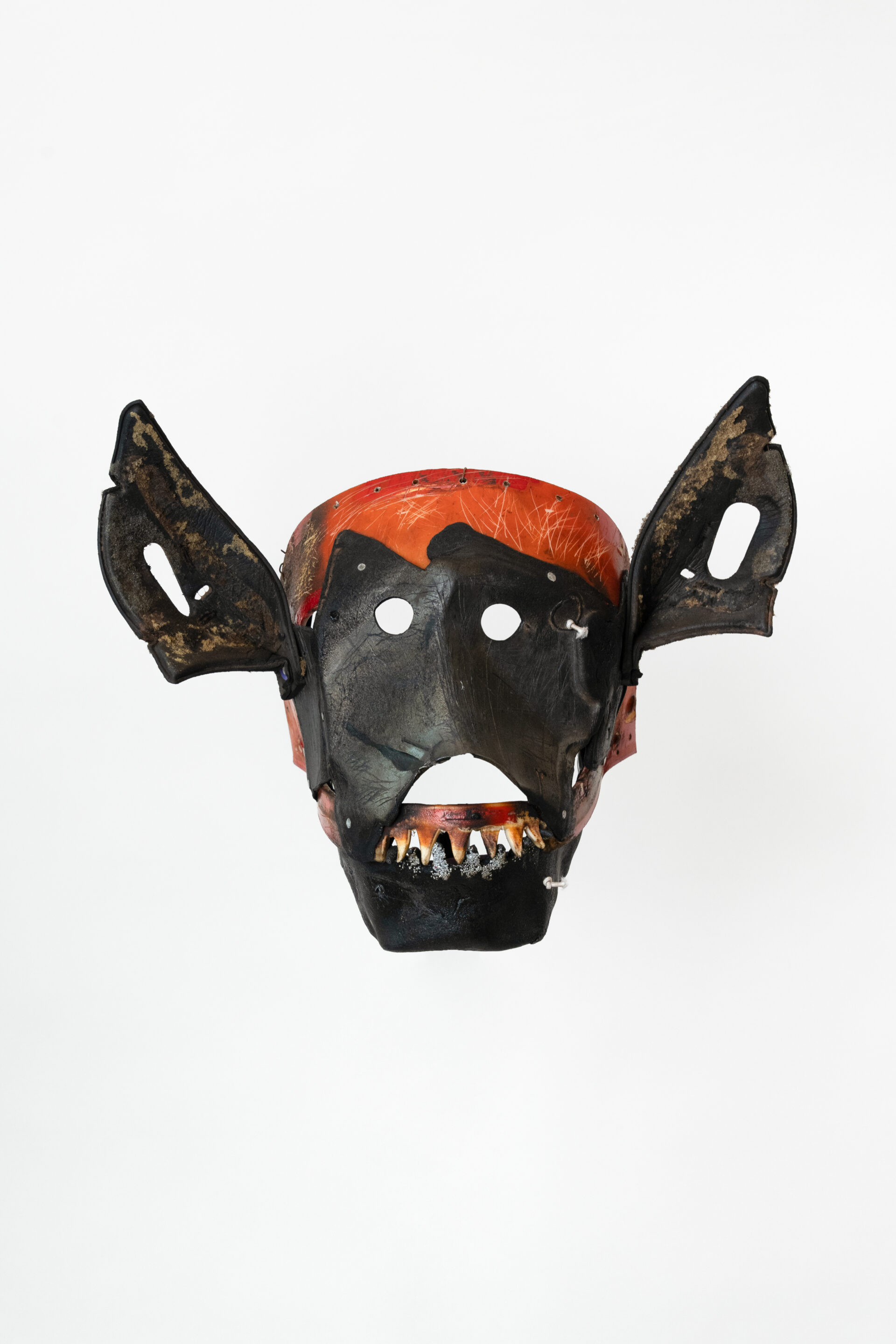 Descrição da imagem: Máscara tridimensional pintada. Vista de frente, a obra é predominantemente preta, com a testa em vermelho. As grandes orelhas são triangulares, em preto e marrom, com algumas áreas desgastadas e a abertura dos ouvidos é ovalada.
Os olhos são pequenos, redondos e vazados. A boca está aberta: a parte superior é arqueada e voltada para baixo, a inferior tem pequenas pontas triangulares. Sobreposto a parte inferior, há uma gengiva com dentes amarelados, voltados para baixo e manchados de preto. Fim da descrição da imagem.
